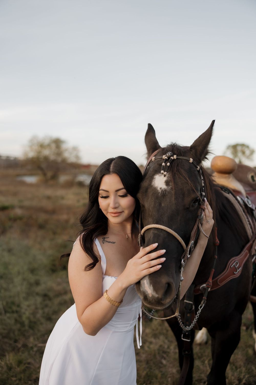 Beautiful brunette woman in white dress gently holds dark brown horse adorned with floral bridle in field.