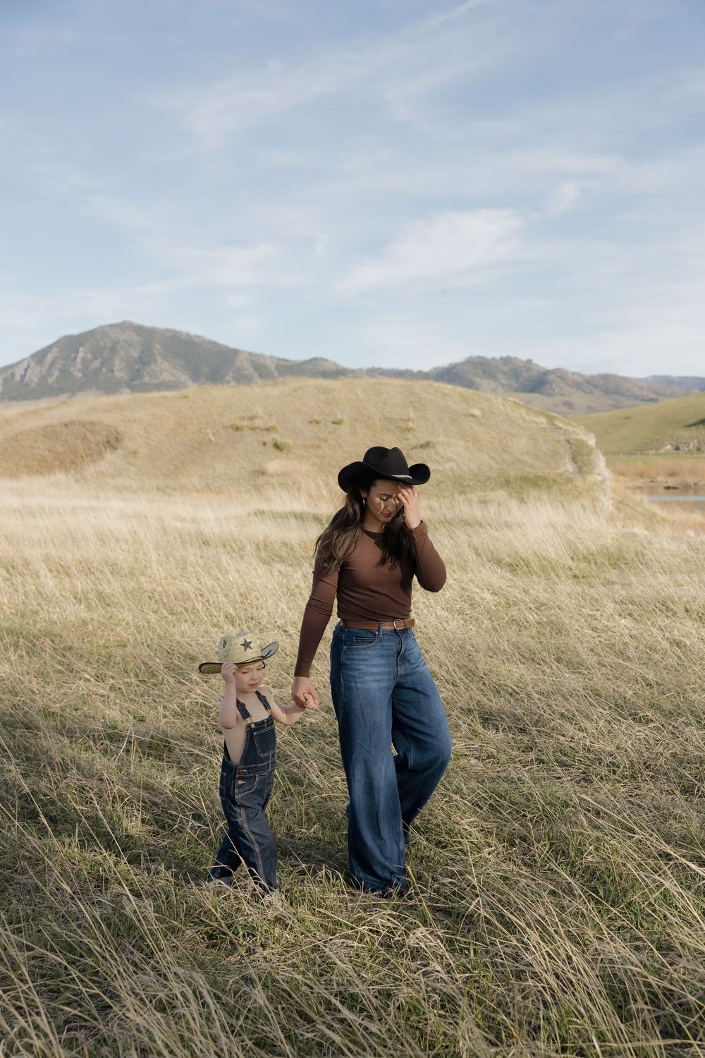 Mother and young son wearing cowboy hats holding hands while walking through tall golden grass.