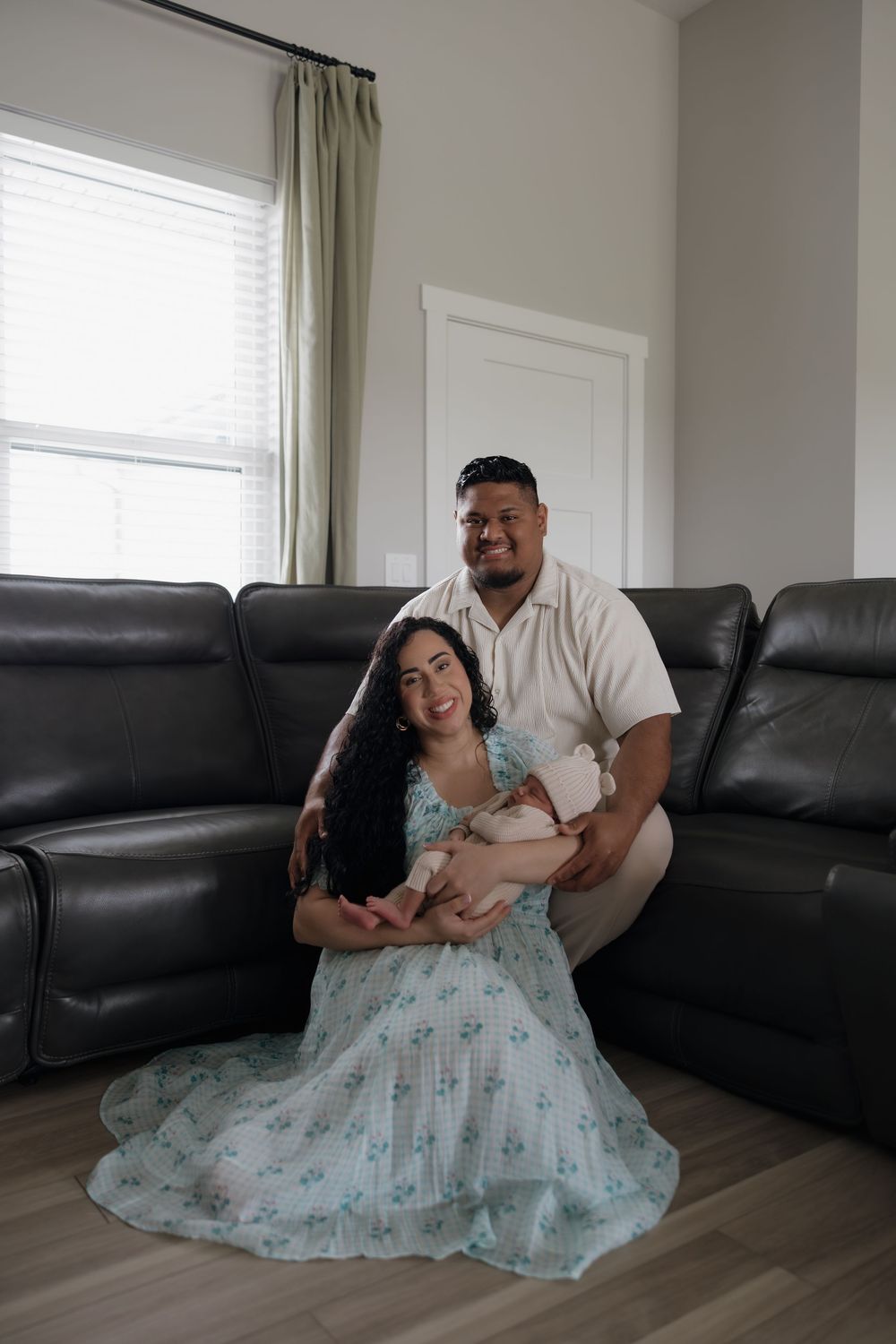 A smiling couple sits on a leather sofa holding their newborn baby in a bright living room.