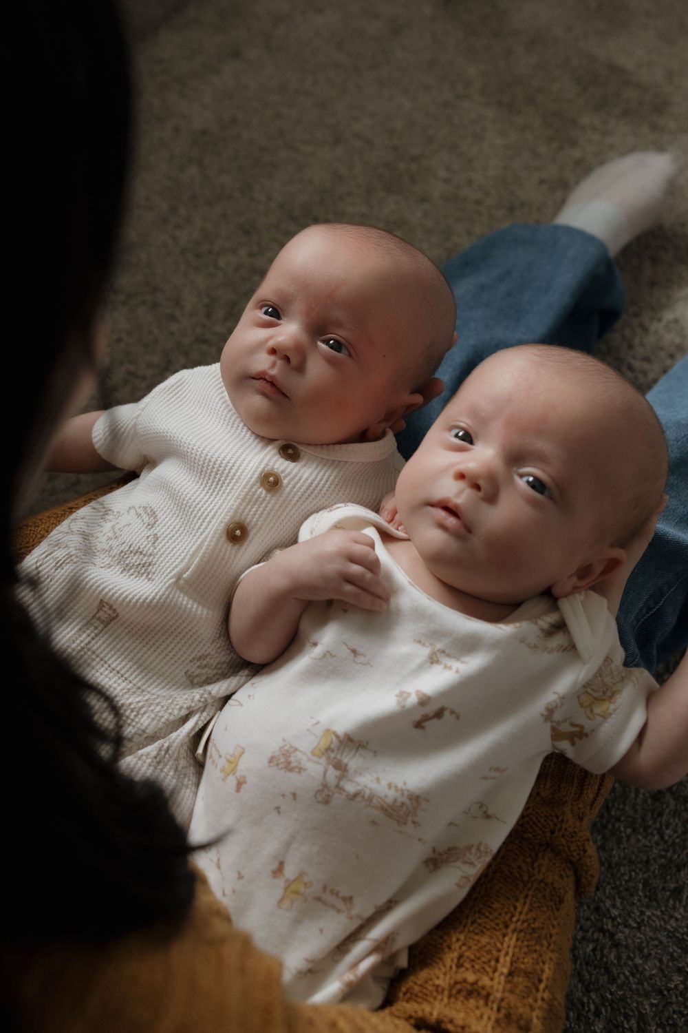 Two newborn twins lying on a carpet, dressed in white onesies, looking up at their mother.