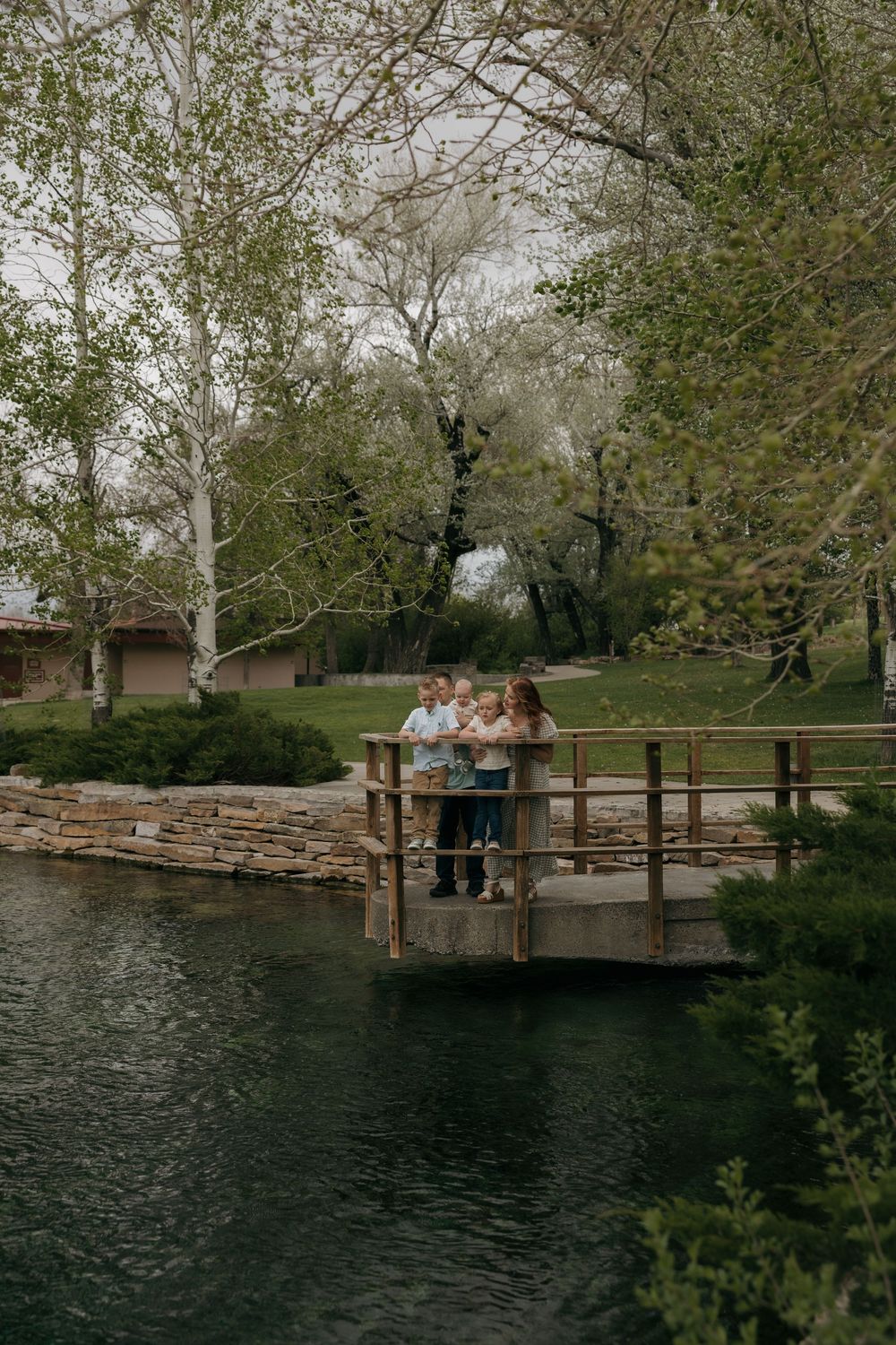 Family with children posing on wooden dock bridge over calm pond in green park.