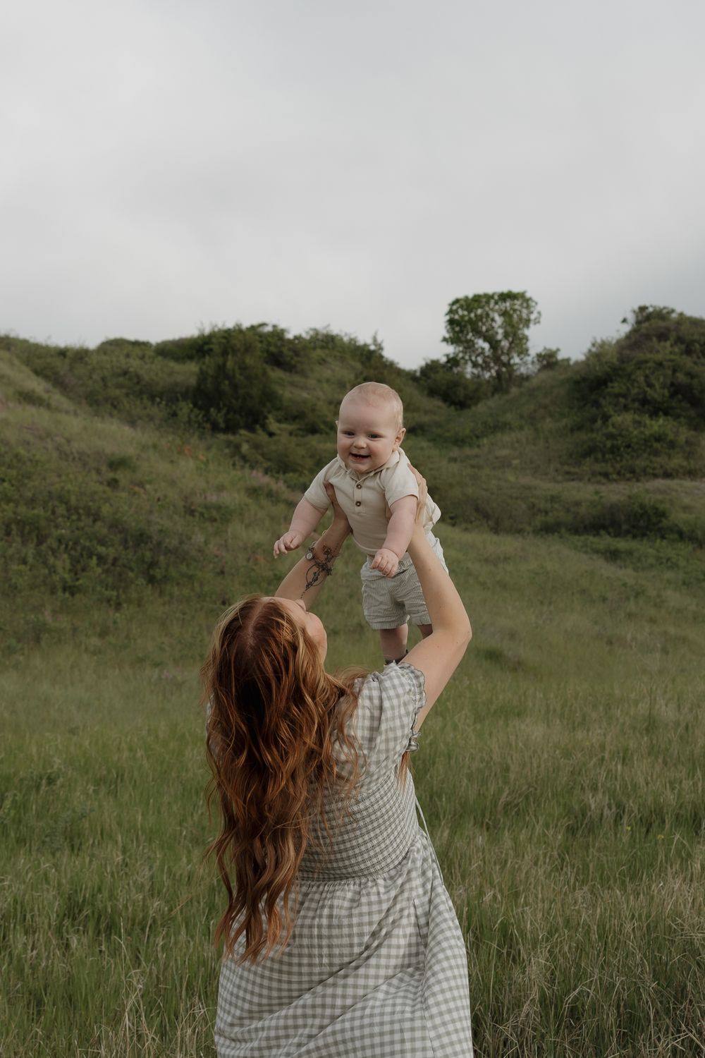 Mother lifting happy laughing baby in air in green grassy meadow on overcast day.