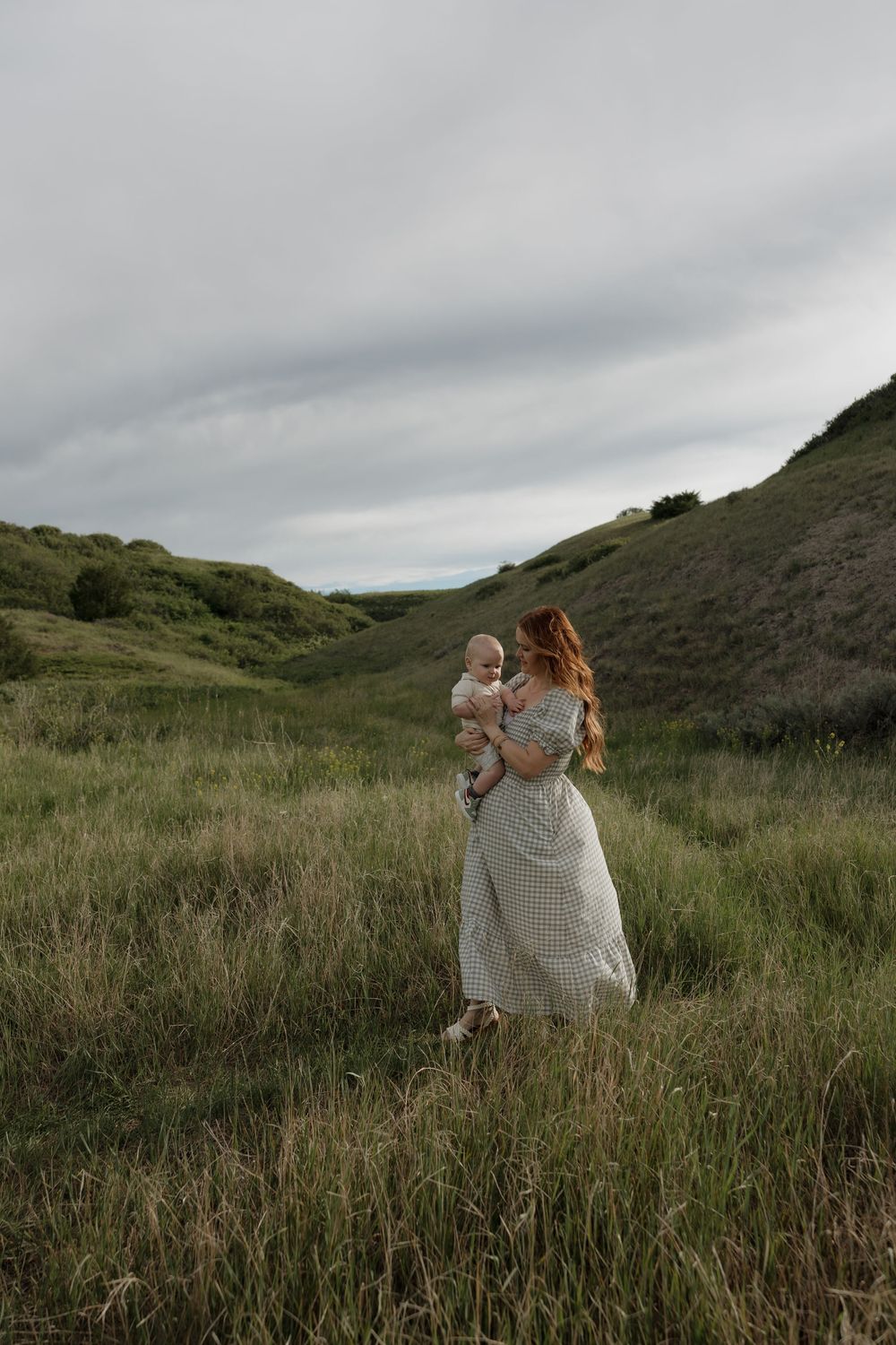 Redhead mother in gingham dress holding baby in green meadow with rolling hills backdrop.