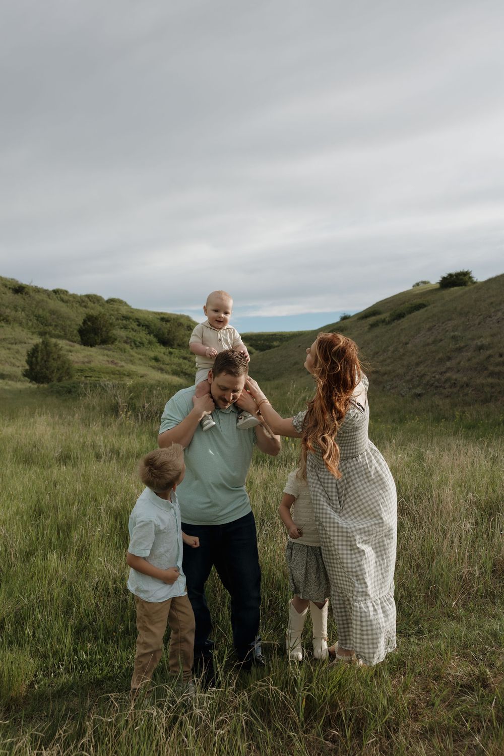 Happy family with three children playing in green meadow with rolling hills on cloudy day.