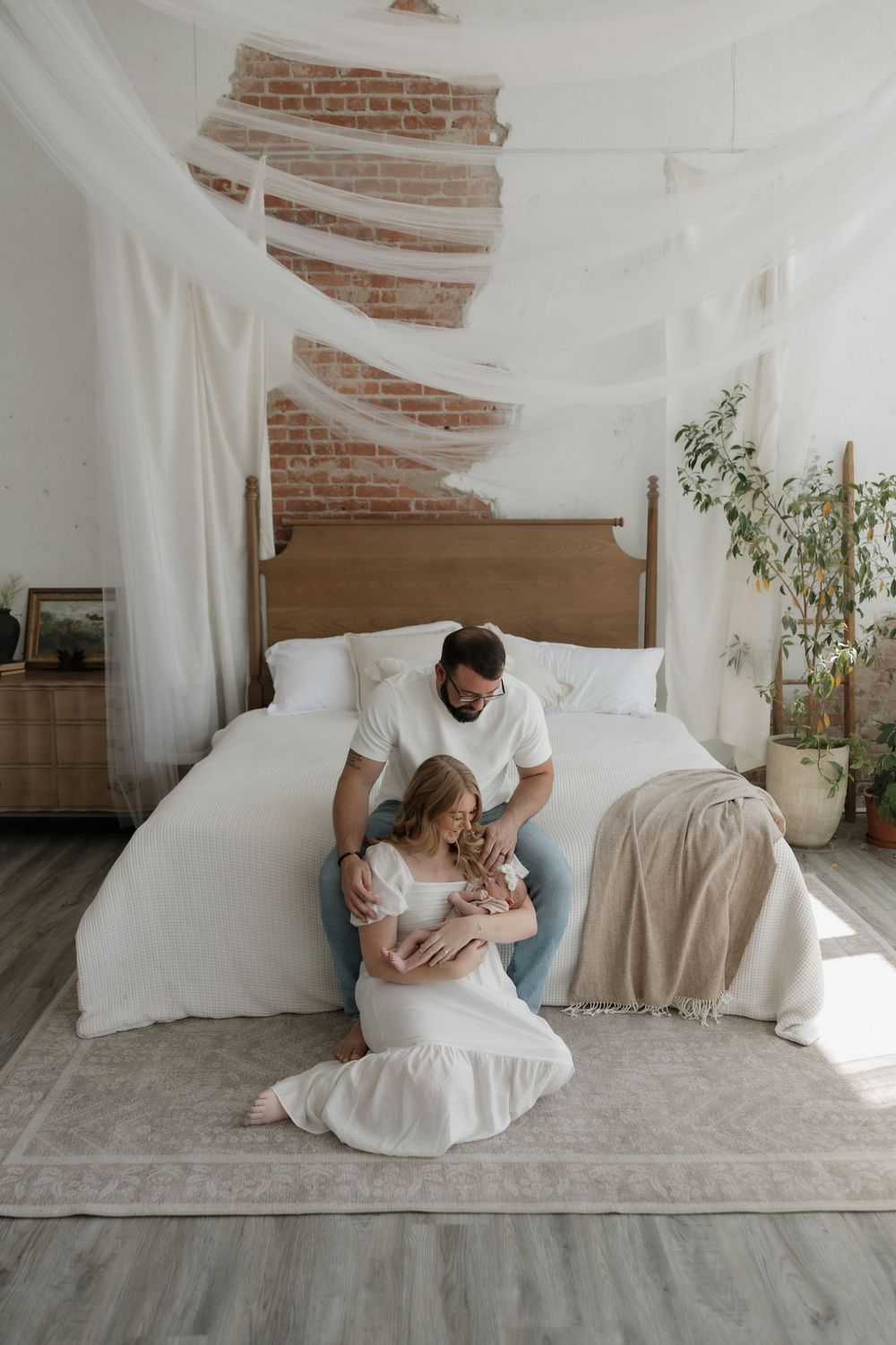 New family with mother in white dress and father in jeans cradling newborn on bedroom floor.