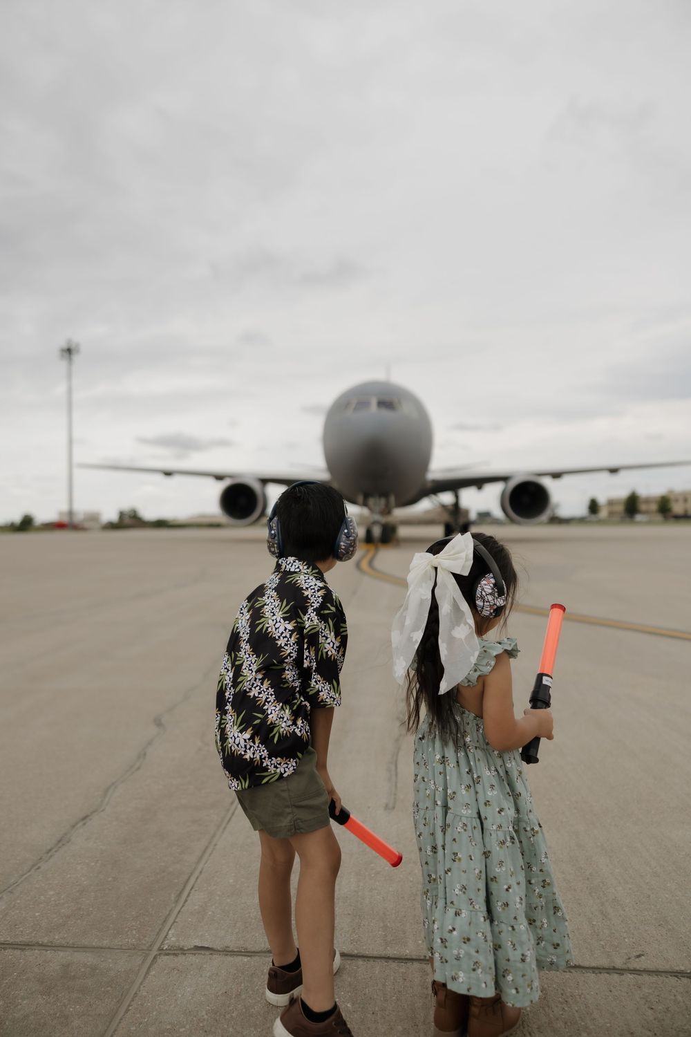 Two children holding light wands watching military aircraft on airport tarmac on overcast day.