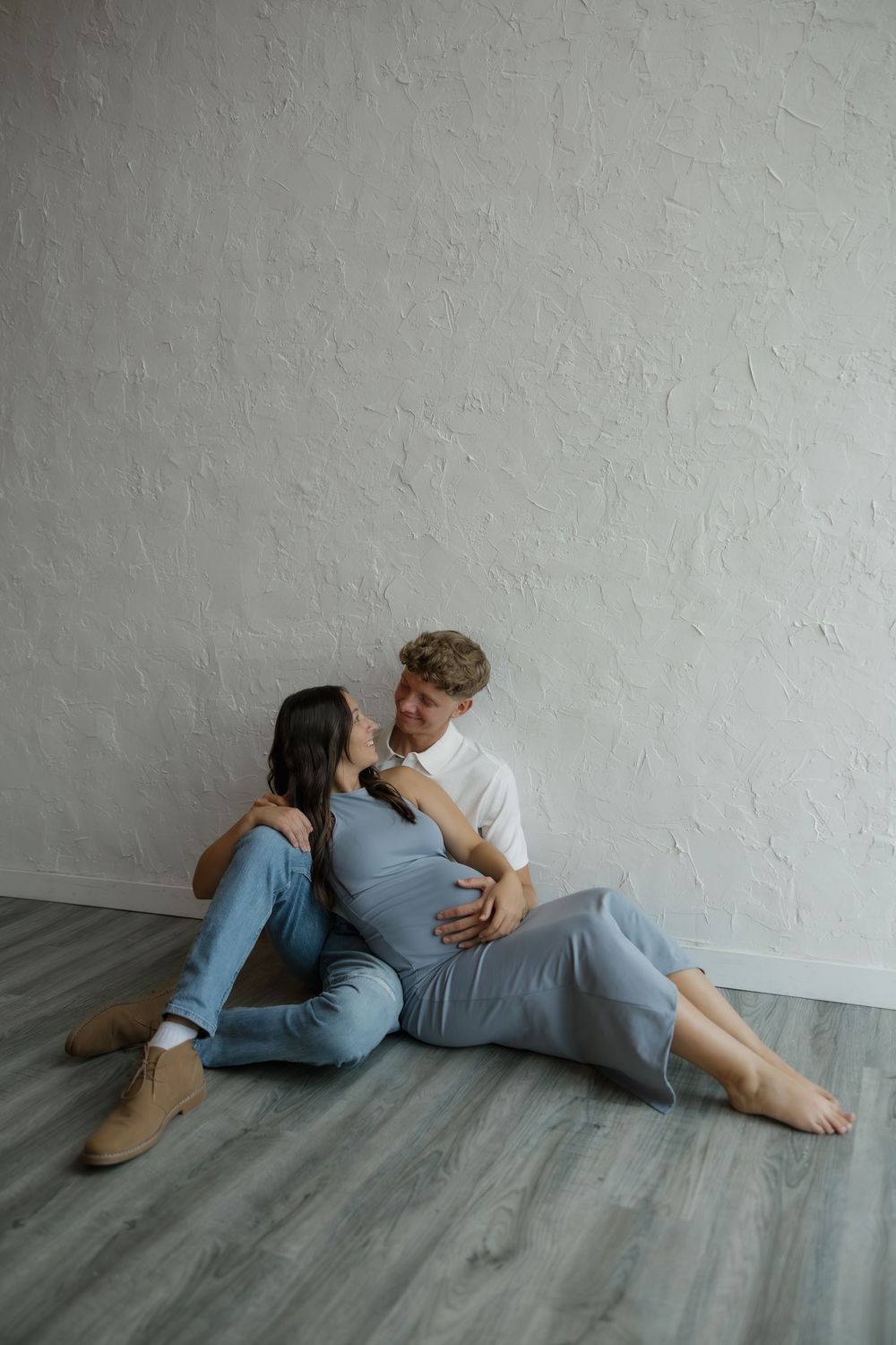 Couple sharing intimate moment on hardwood floor, man embracing pregnant woman in blue dress against white wall.