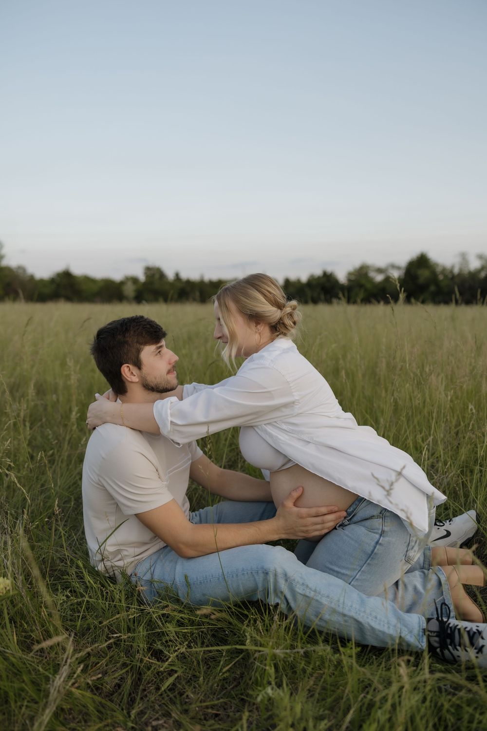 Pregnant couple sitting in lush green field, woman leaning toward man in tender romantic moment outdoors.