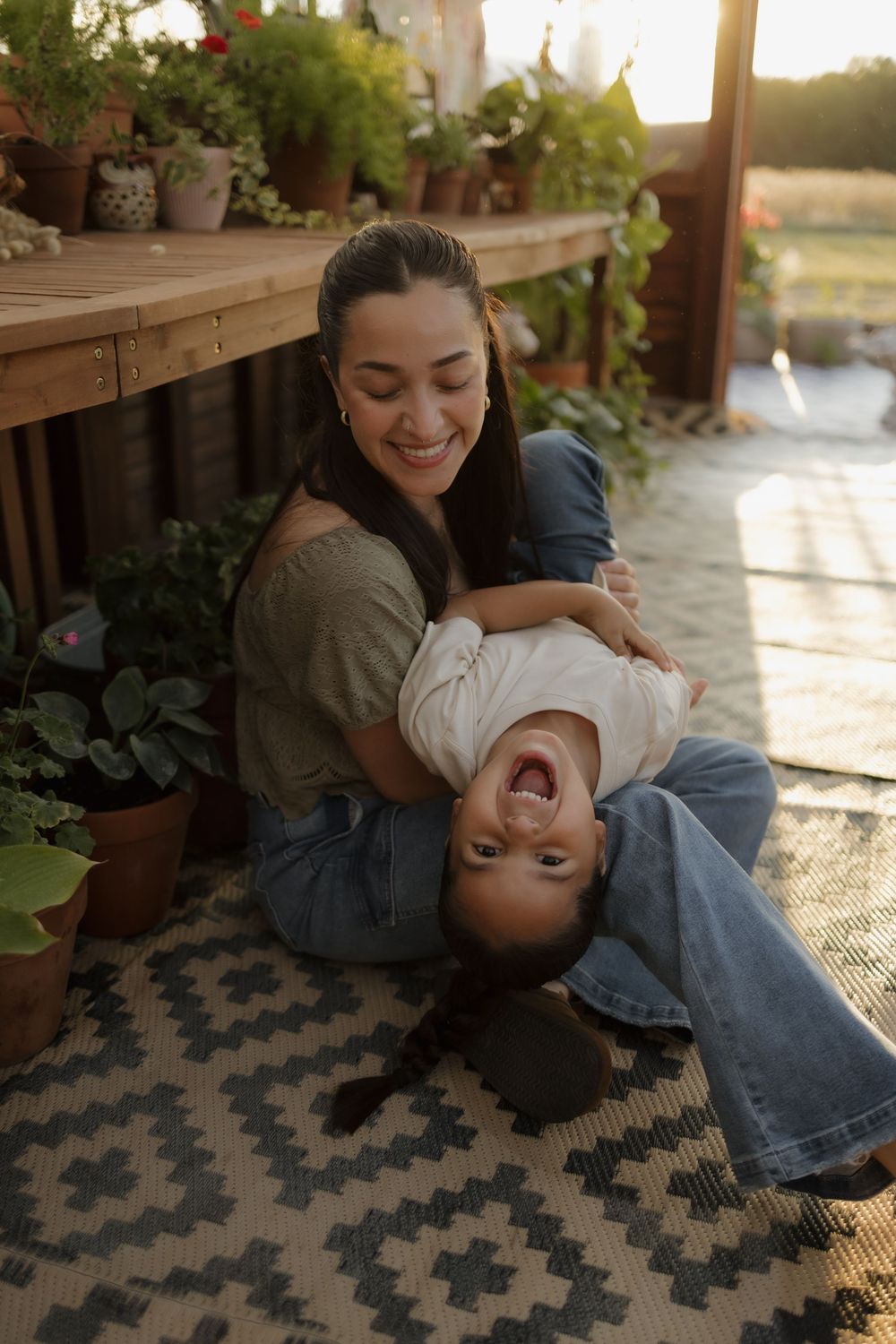 Smiling mother playfully holding laughing child upside down on porch surrounded by potted plants.