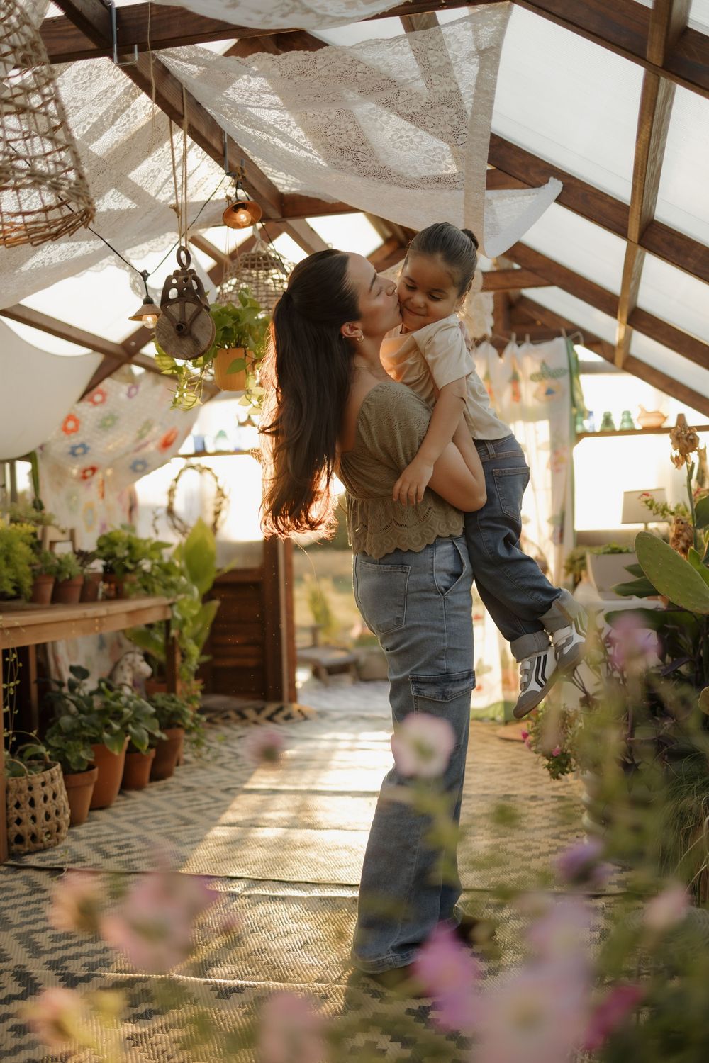 Mother kissing and lifting young child inside a sunlit greenhouse filled with lush green plants.