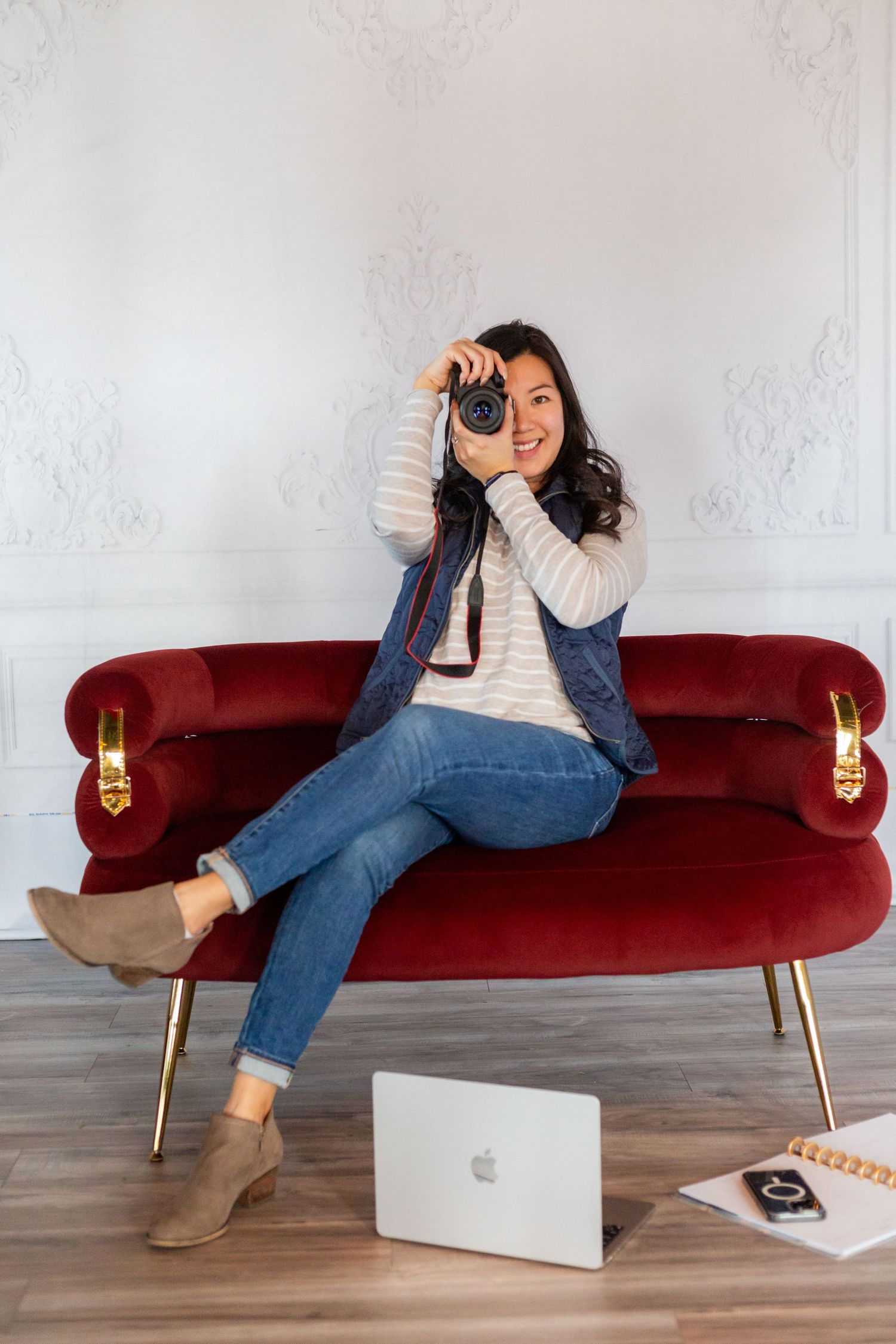 Young woman sitting on hardwood floor smiling at laptop with camera nearby in bright workspace.