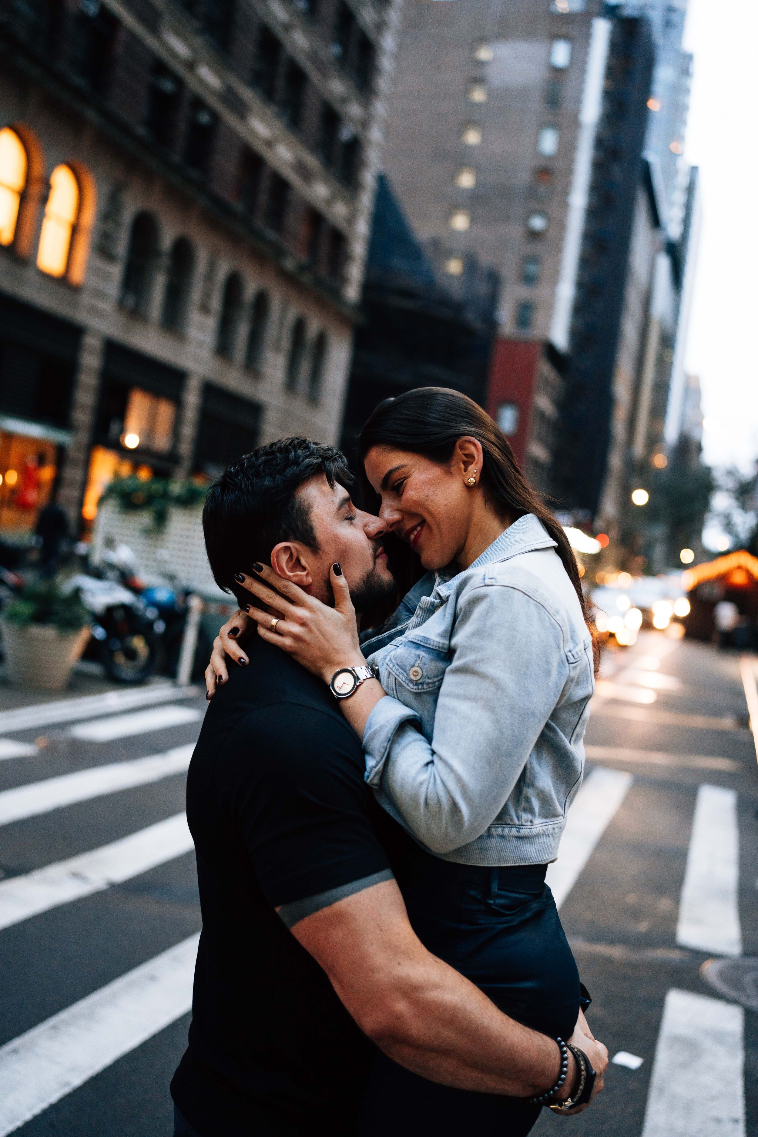 A couple embraces tenderly on a city crosswalk at dusk with urban buildings in the background.