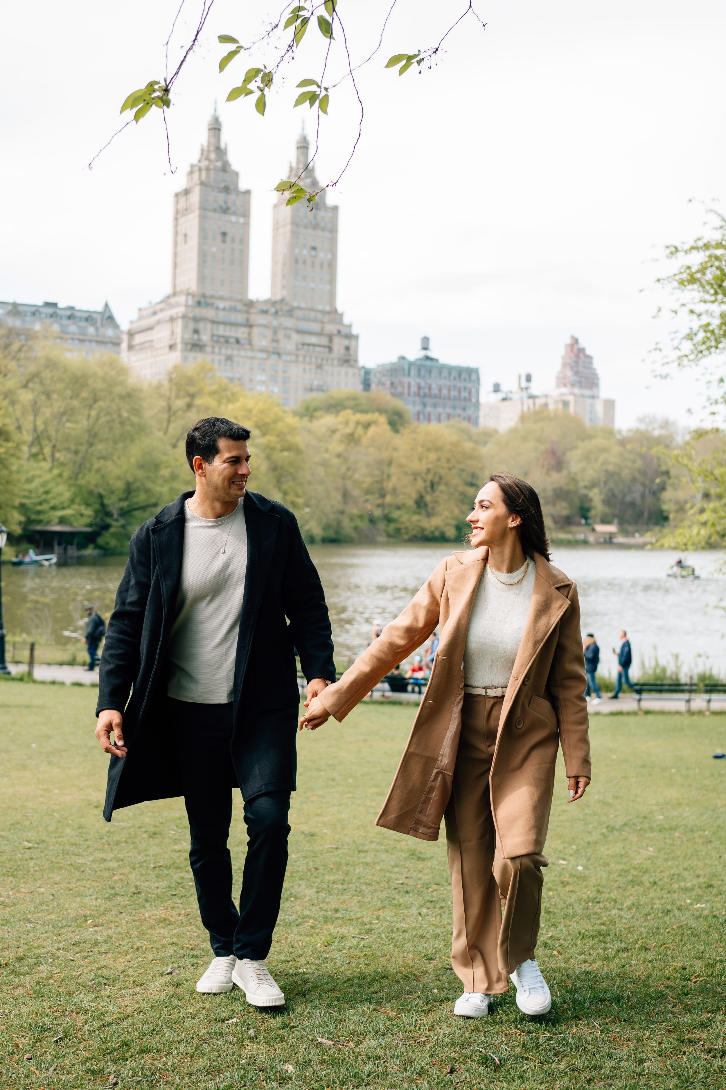 Couple walking hand in hand through Central Park with iconic NYC buildings in background.