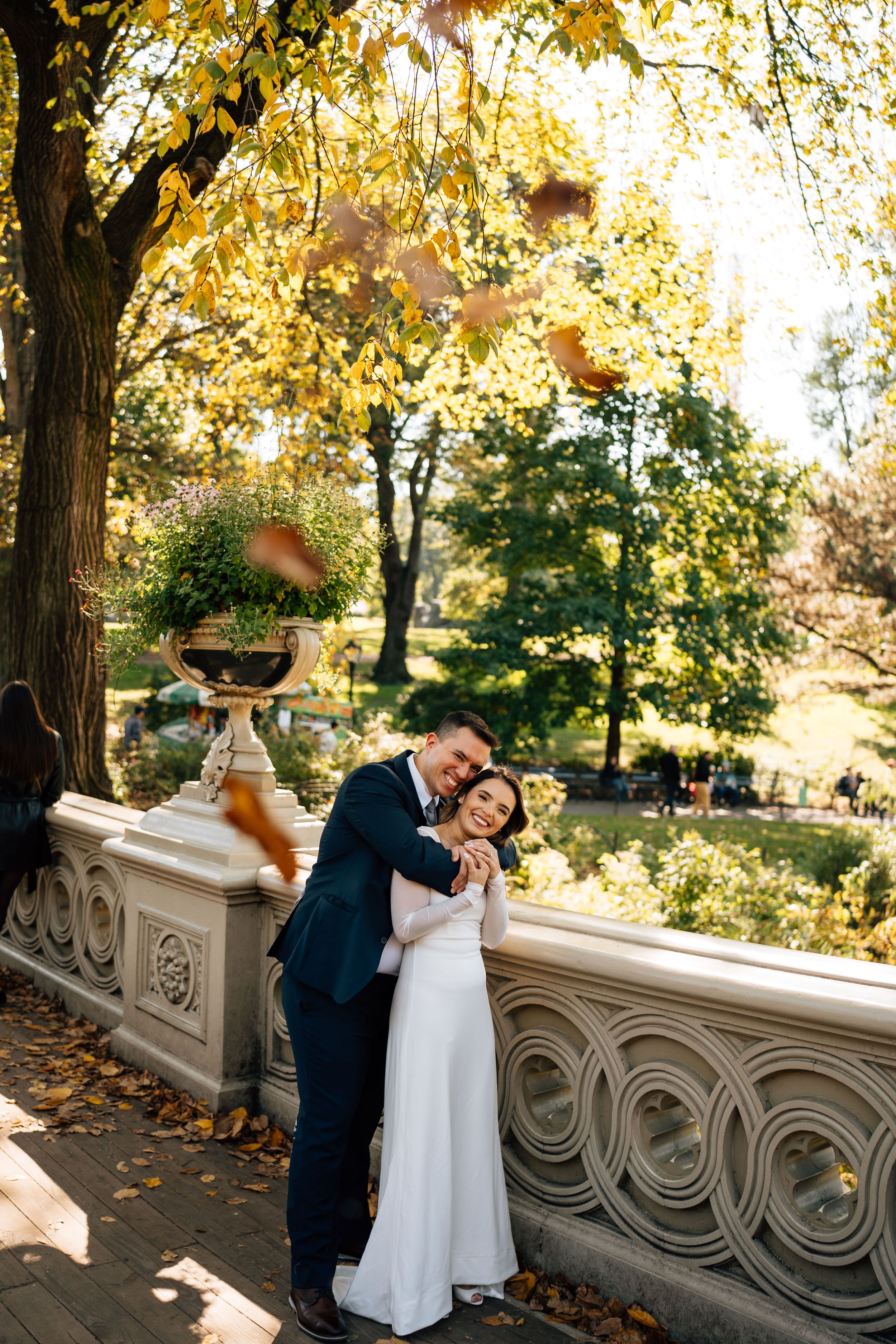 A romantic wedding couple embraces on an ornate stone bridge surrounded by golden autumn foliage.