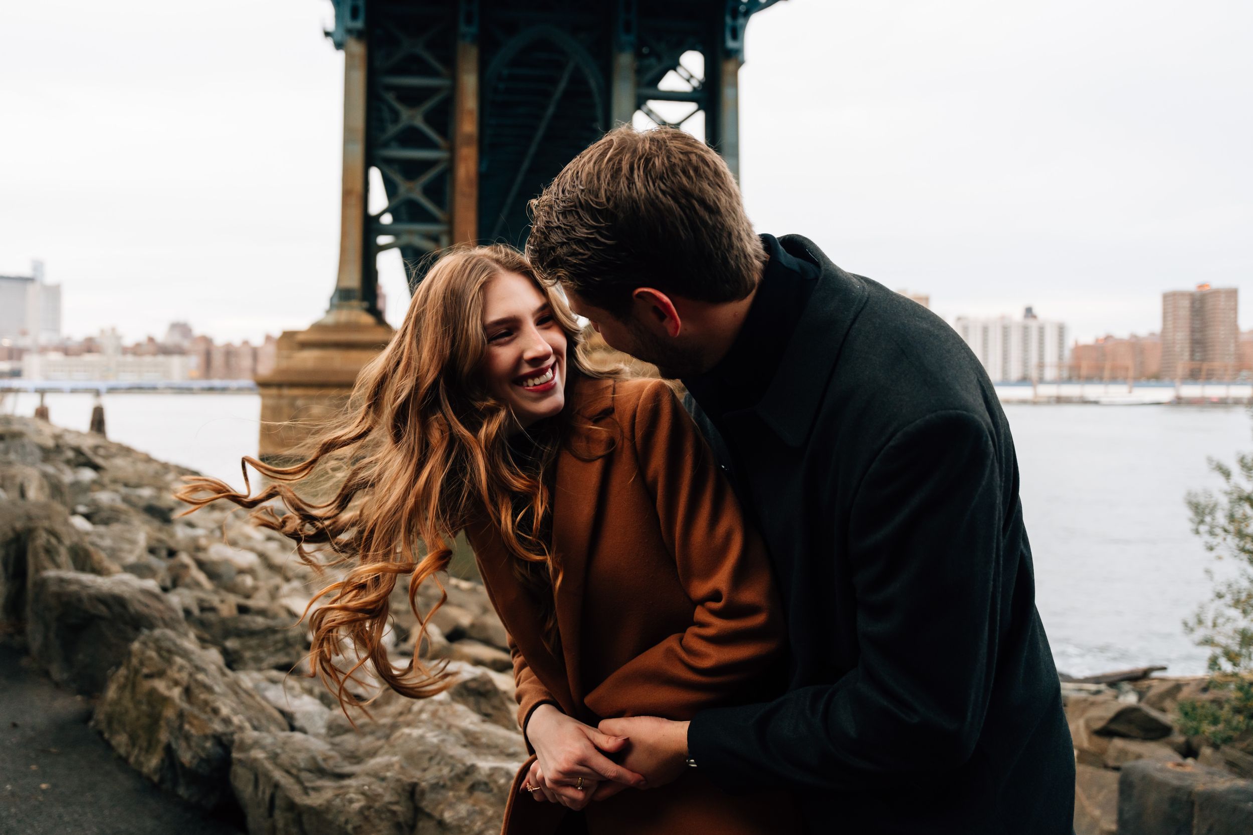 A couple shares a joyful moment together by the waterfront with city buildings visible in the distance.