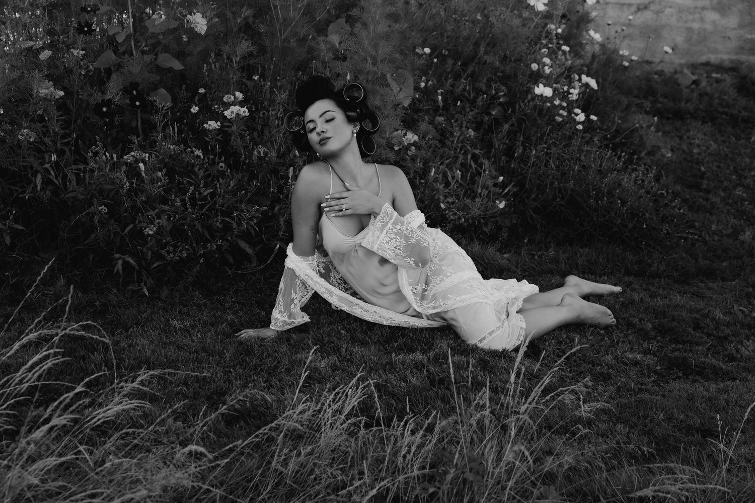 Black and white photo of a woman reclining gracefully on grass in a white dress beside garden flowers.