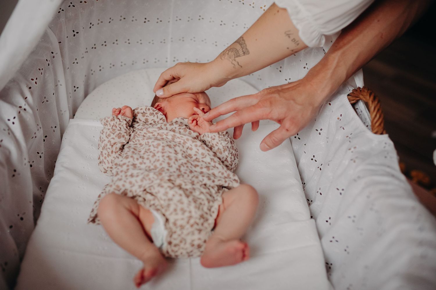 Mother gently touching newborn baby lying in white wicker bassinet wearing floral romper.