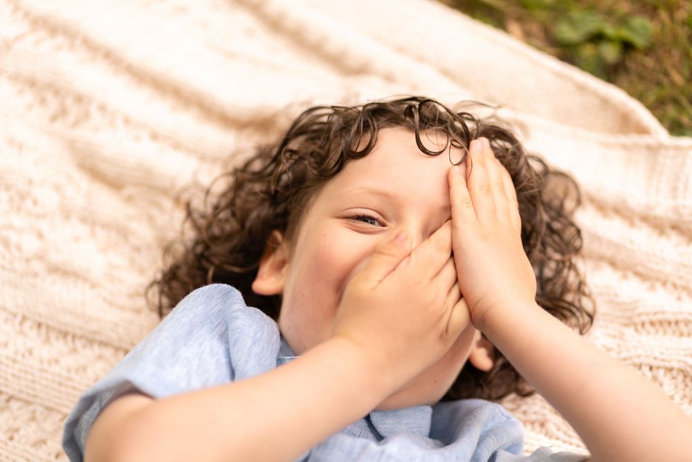 Curly-haired child lying on a blanket outdoors, laughing and covering their face with both hands.