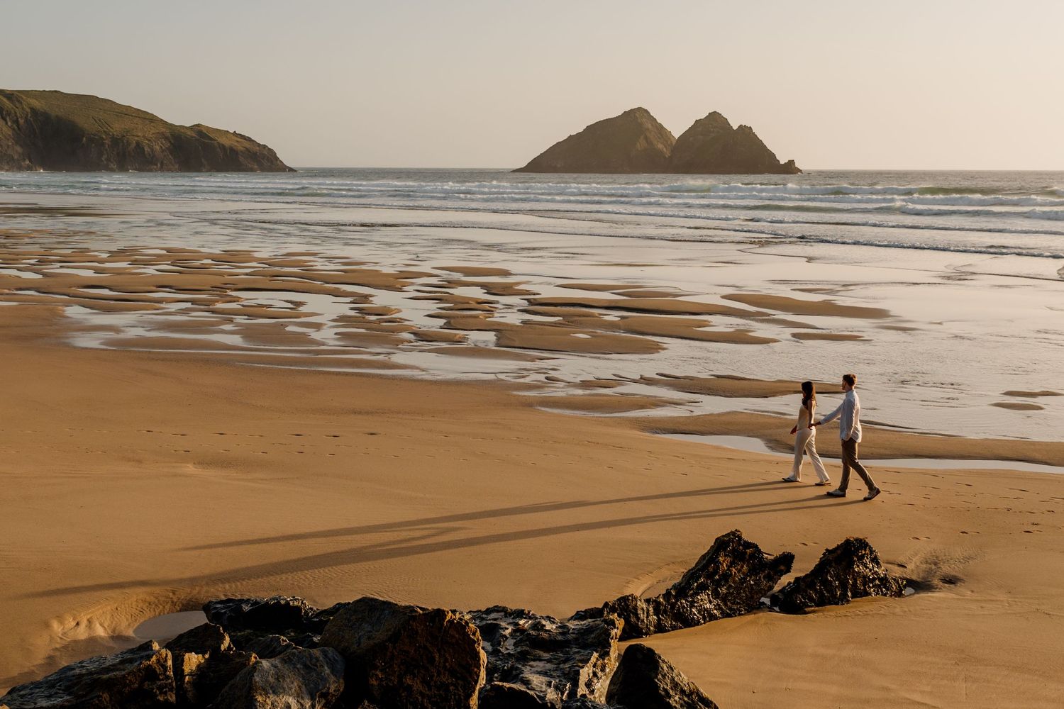 Couple walking hand in hand on Holywell Beach at sunset with rocky islands in the background.