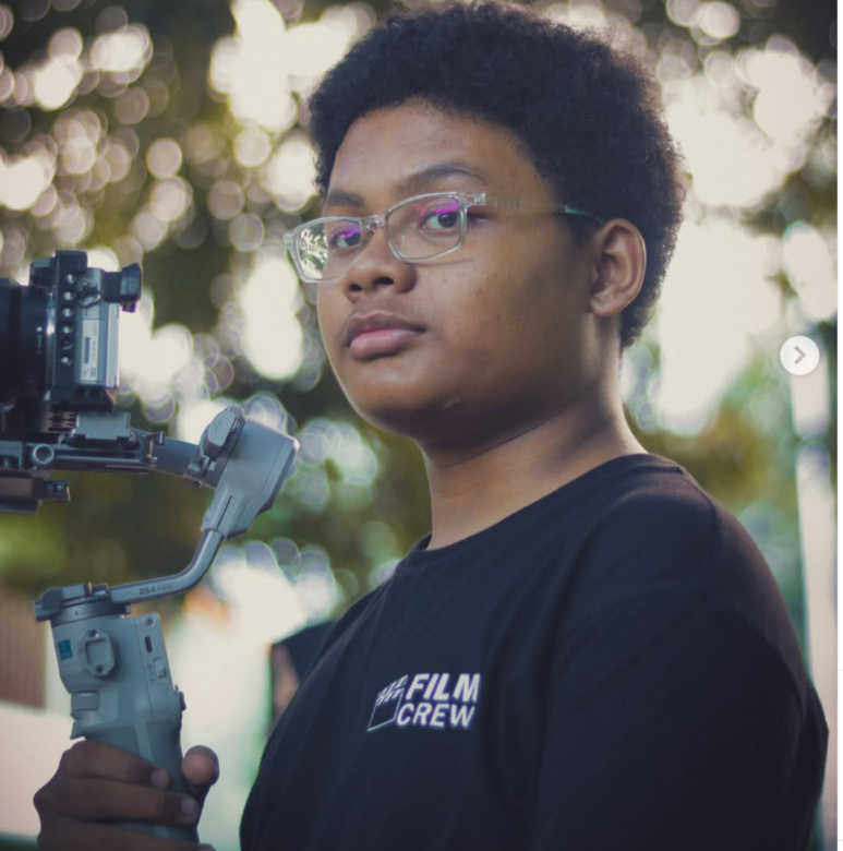 Young filmmaker holding a camera gimbal stabilizer outdoors, wearing a black Film Crew shirt and glasses.