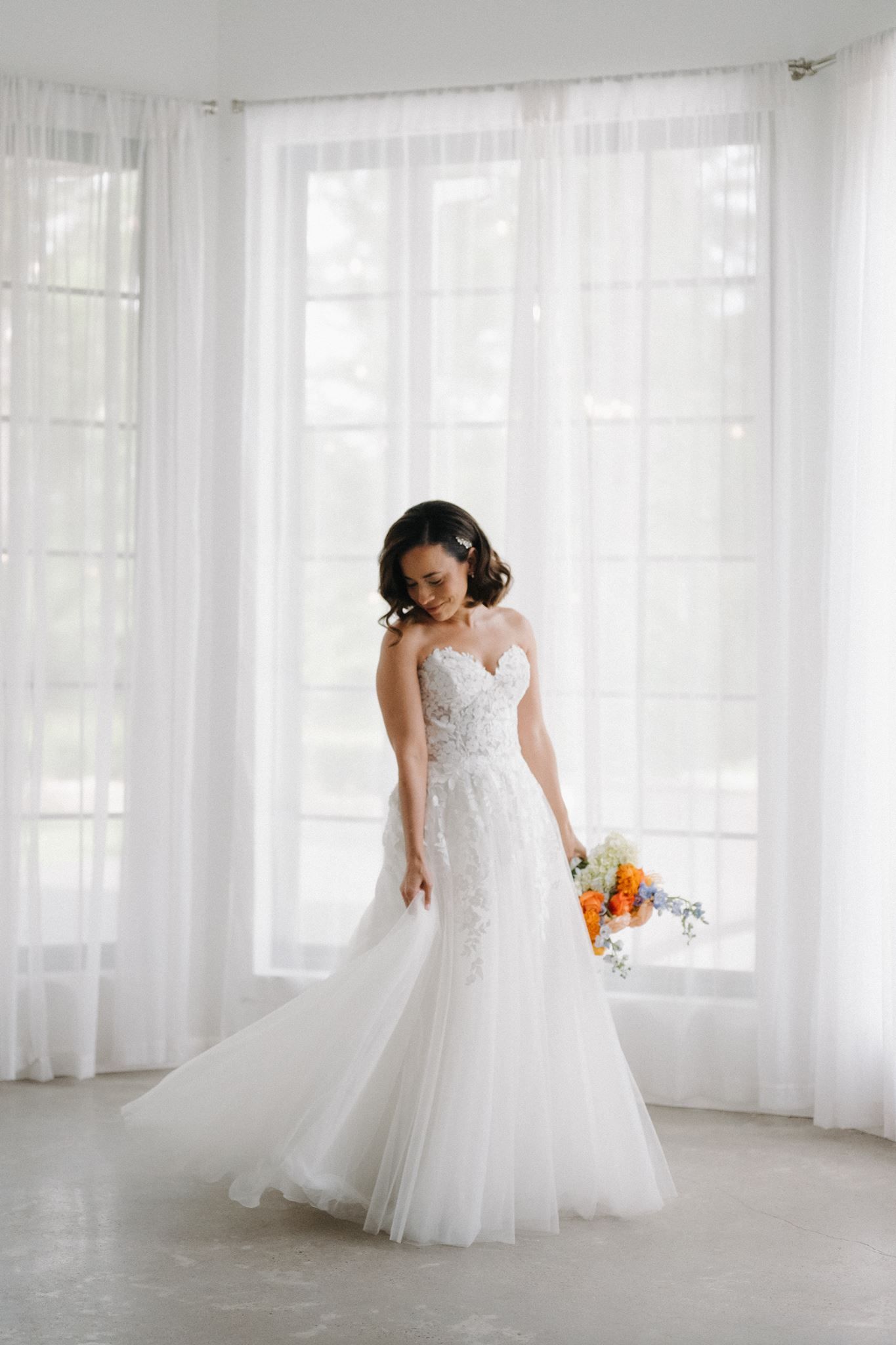 Bride in strapless lace mermaid wedding gown holding colorful bouquet standing before large white curtained windows.