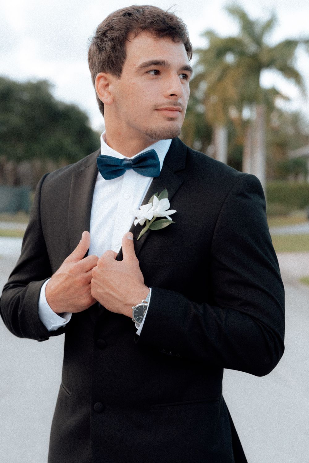 Elegant groom in black tuxedo with teal bow tie and white boutonniere adjusts jacket outdoors.