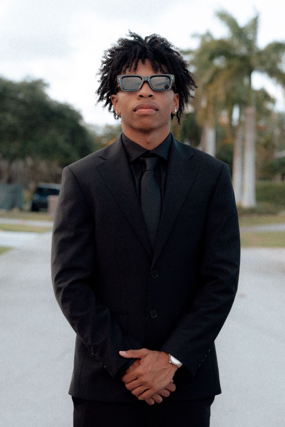 Stylish young man in black suit and sunglasses poses confidently with palm trees in the background.