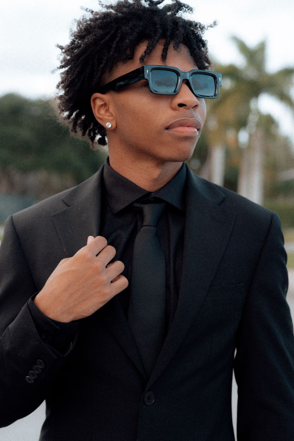Stylish young man in all-black suit and teal sunglasses poses confidently with palm trees behind.