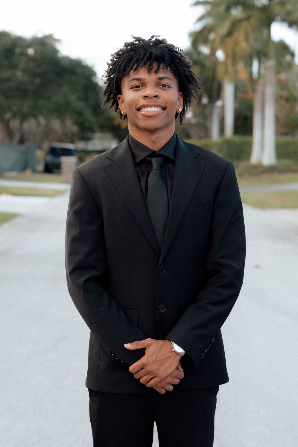 Smiling young Black man in black suit and tie posing outdoors with palm trees in background.