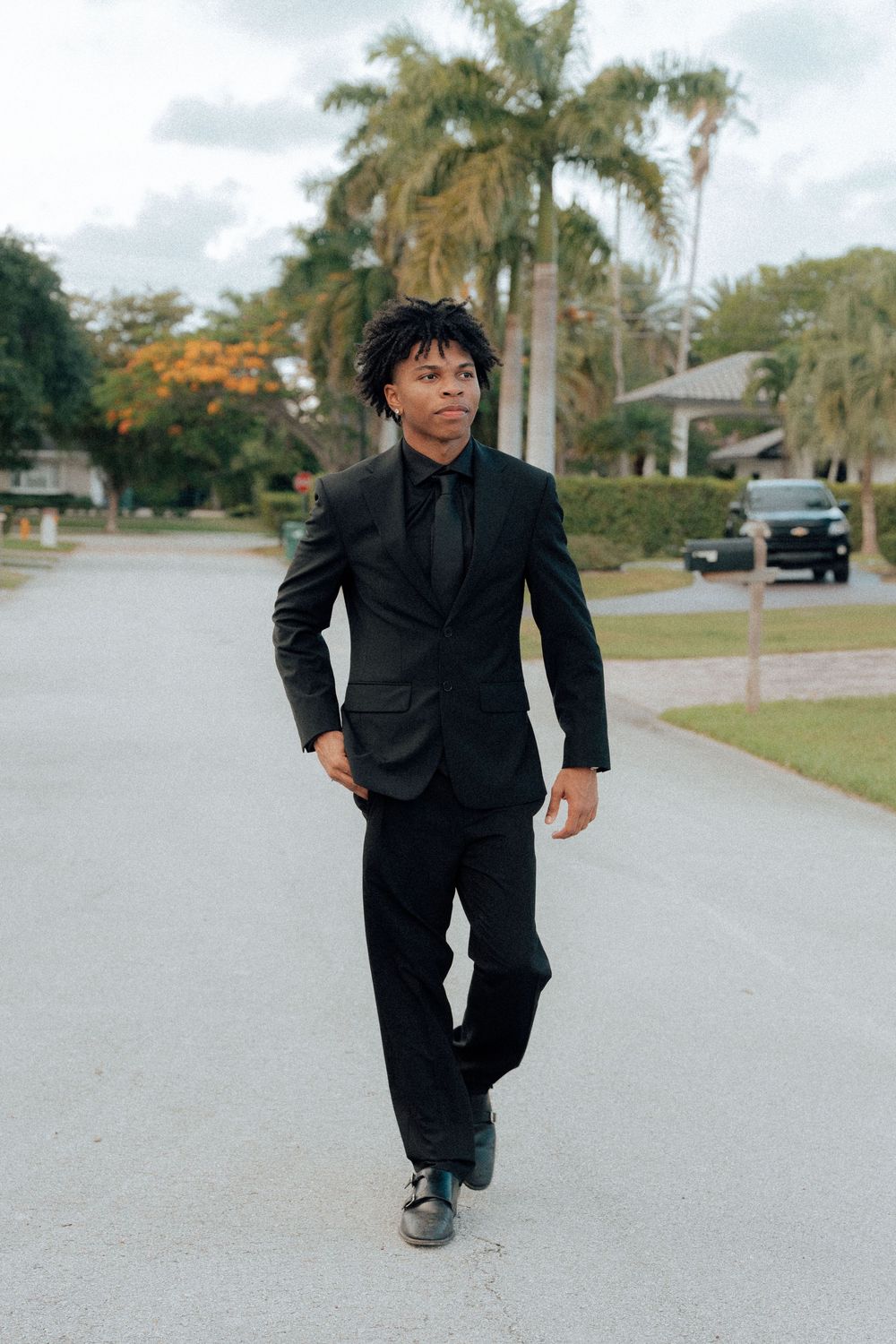 Young man in black suit walks confidently down suburban street lined with palm trees.