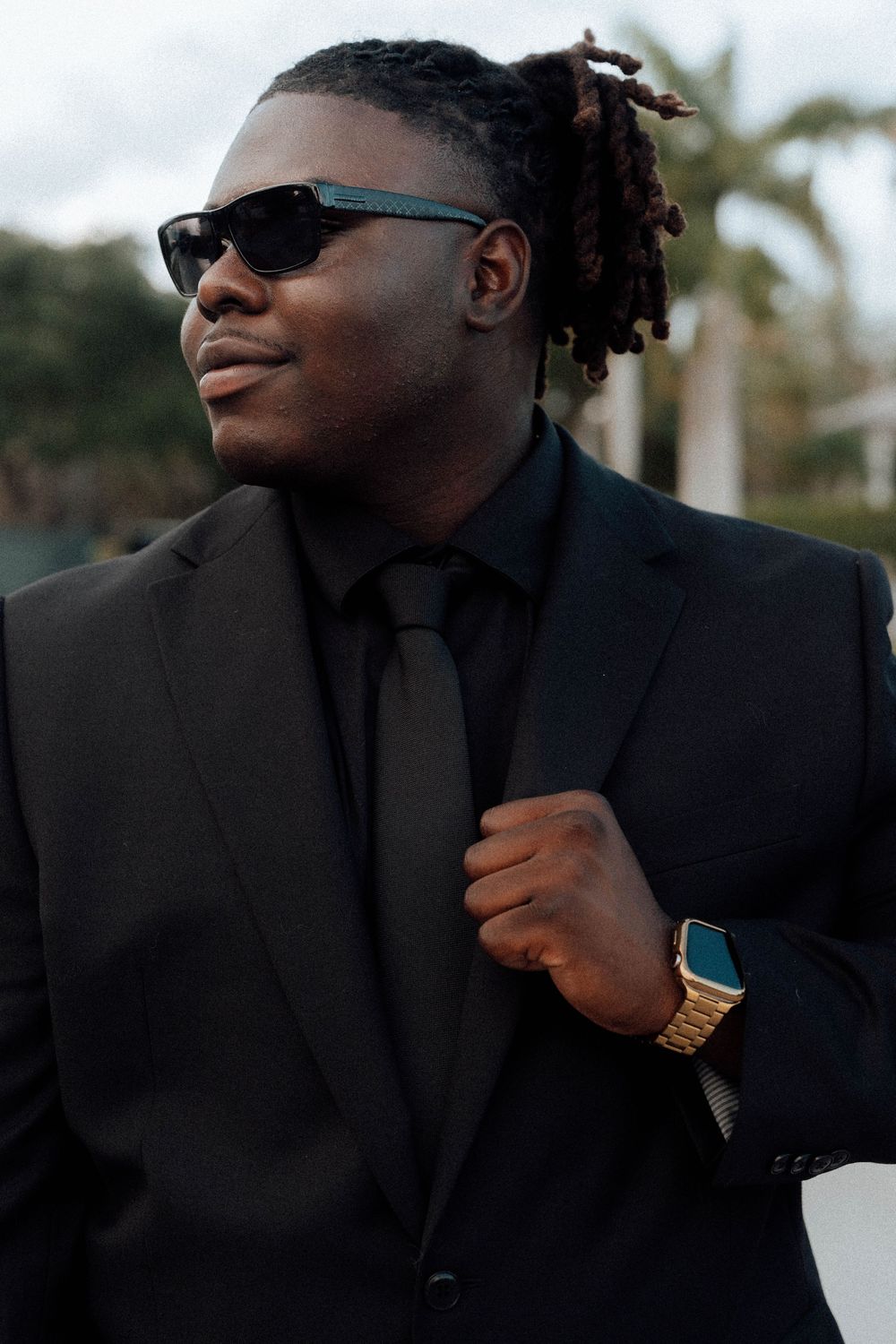 Stylish Black man in black suit and tie wearing sunglasses and gold watch with dreadlocks outdoors.