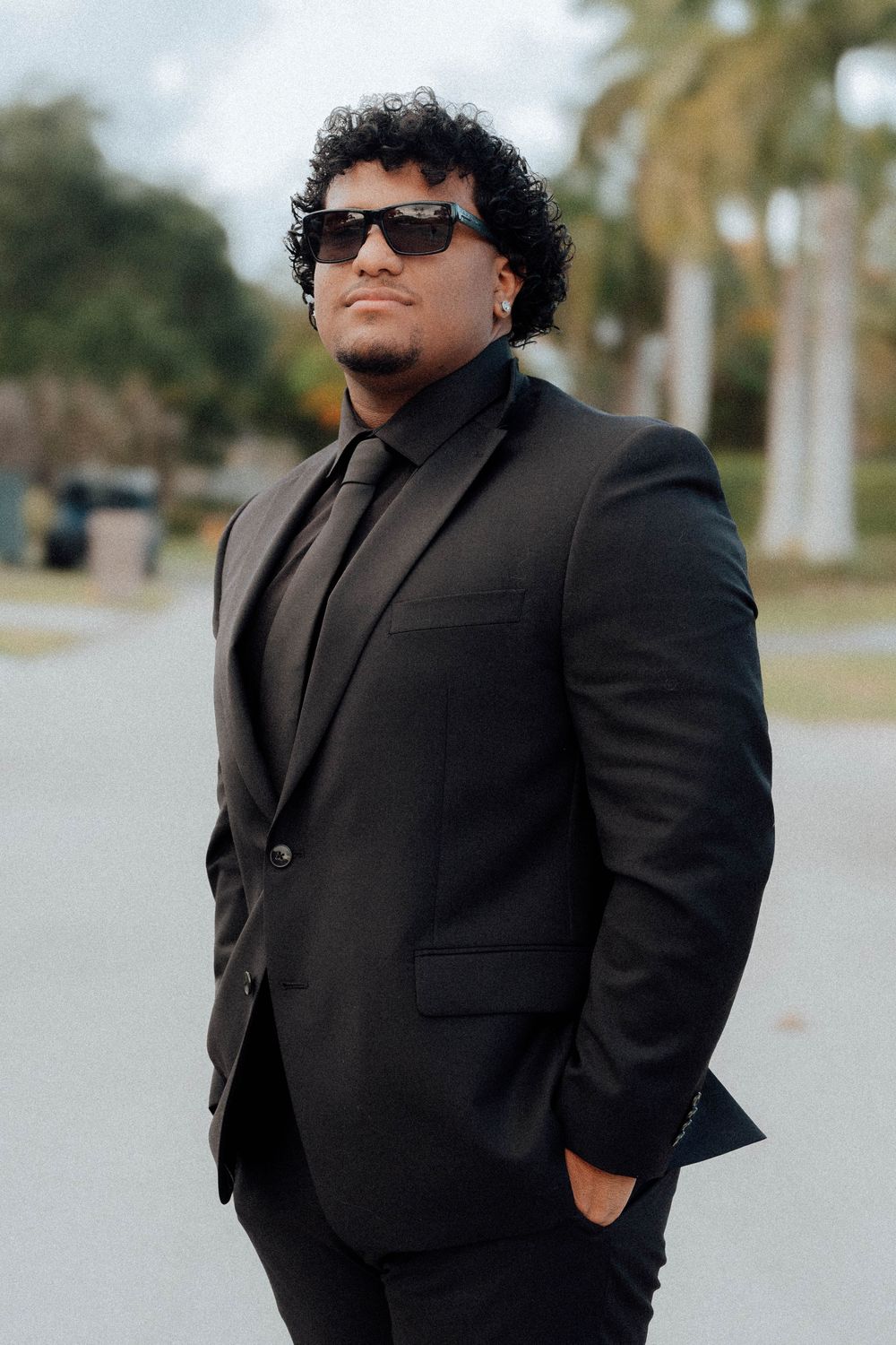 Confident man in black suit and sunglasses poses outdoors with palm trees in background.