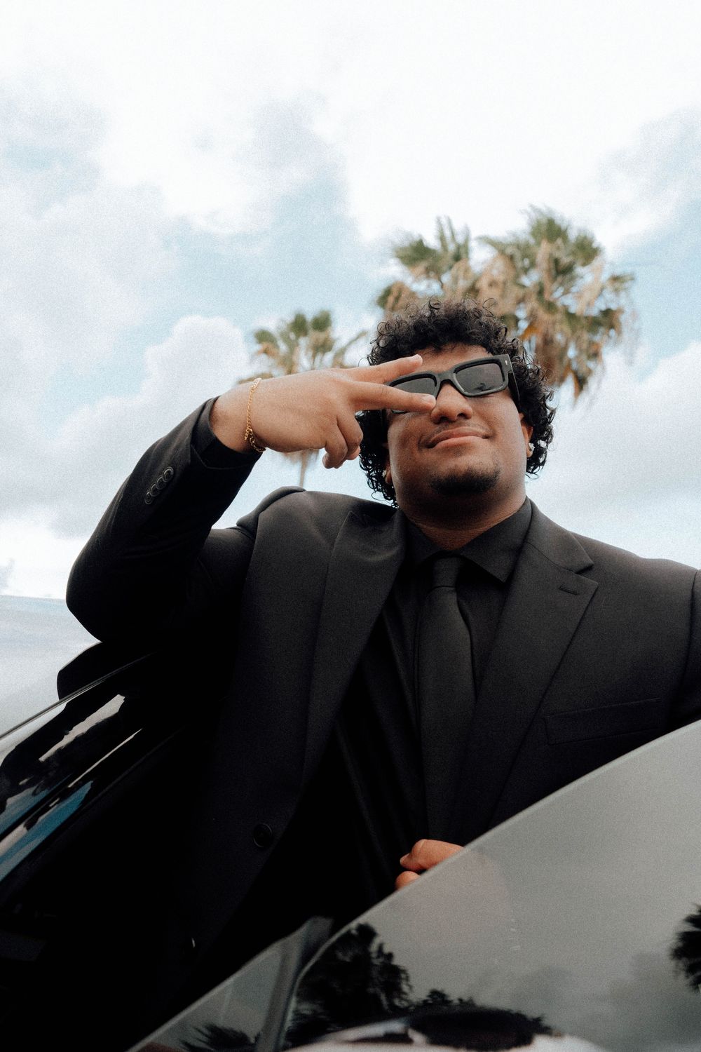 Stylish young man in black suit and sunglasses posing confidently beside a black car with palm trees behind.