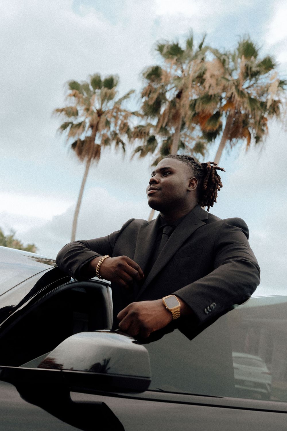 Young Black man in black suit leans on luxury car door with palm trees in background.