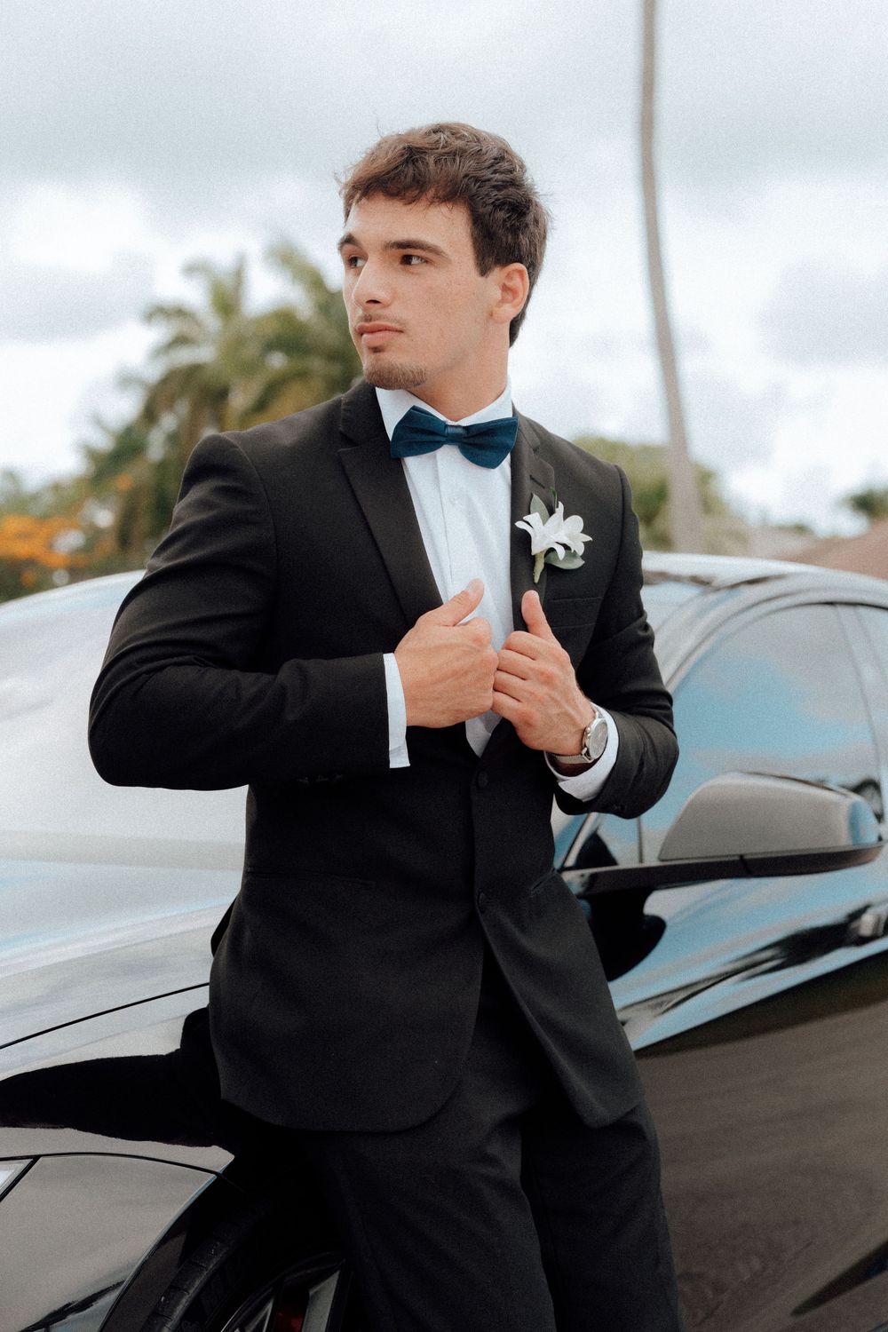 A young man in a black tuxedo with a blue bow tie and boutonniere leaning against a black car outdoors.