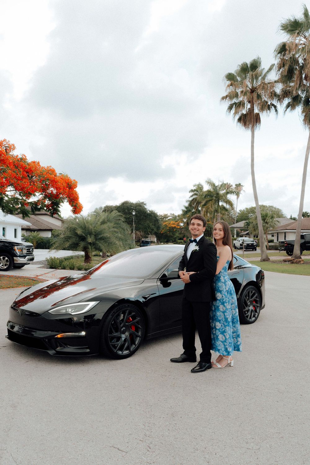 Prom couple in formal attire posing in front of black Tesla with palm trees.