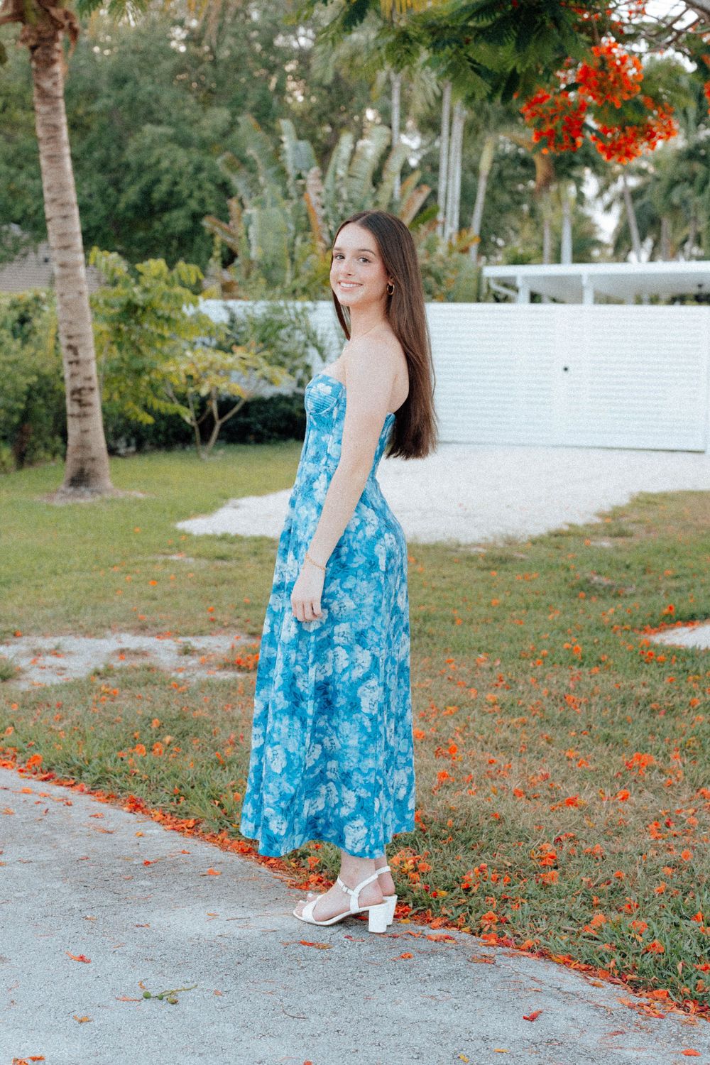 Young woman in blue floral maxi dress and white heels posing outdoors near palm trees.