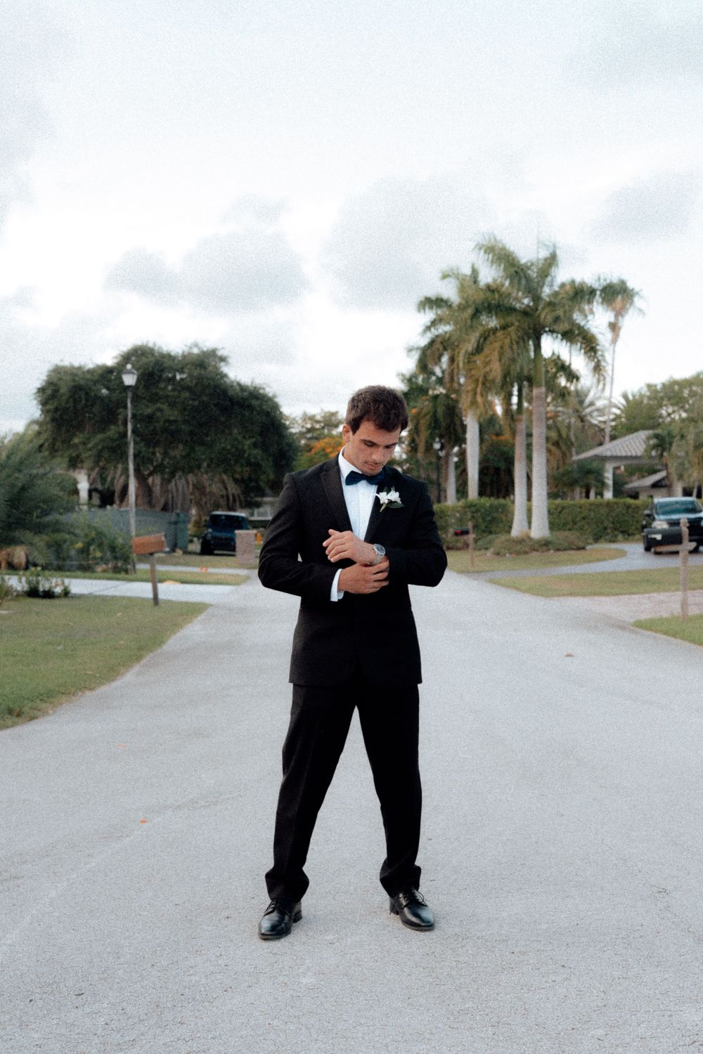 Young man in black tuxedo with bow tie adjusts his cufflinks on a palm tree-lined driveway.