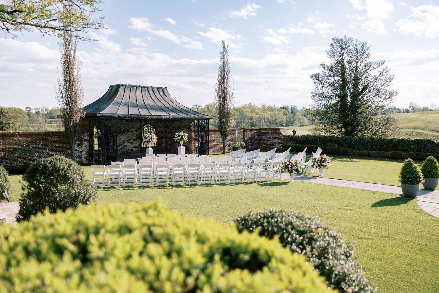 Elegant outdoor wedding ceremony setup with white chairs facing a classic gazebo surrounded by manicured gardens.