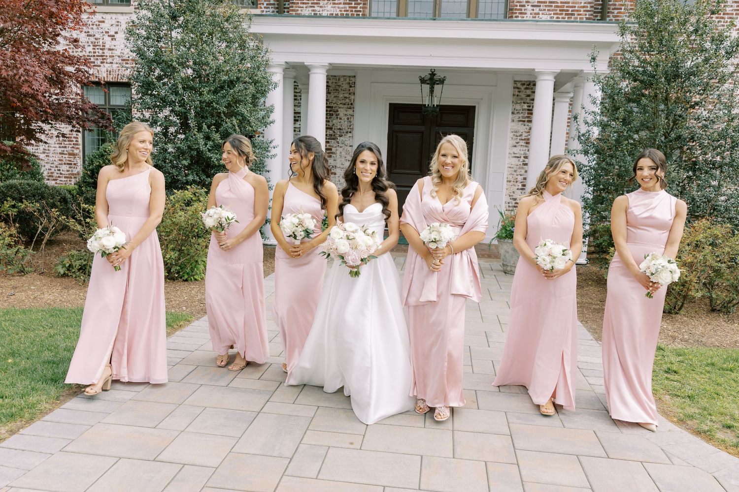 Bride in white gown poses with six bridesmaids in blush pink dresses holding white bouquets outside elegant venue.