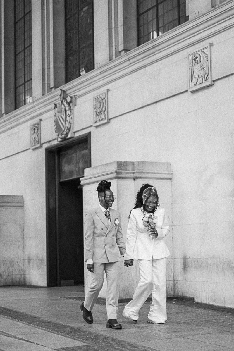 Two brides in white suits hold hands walking outside a grand building on their wedding day.