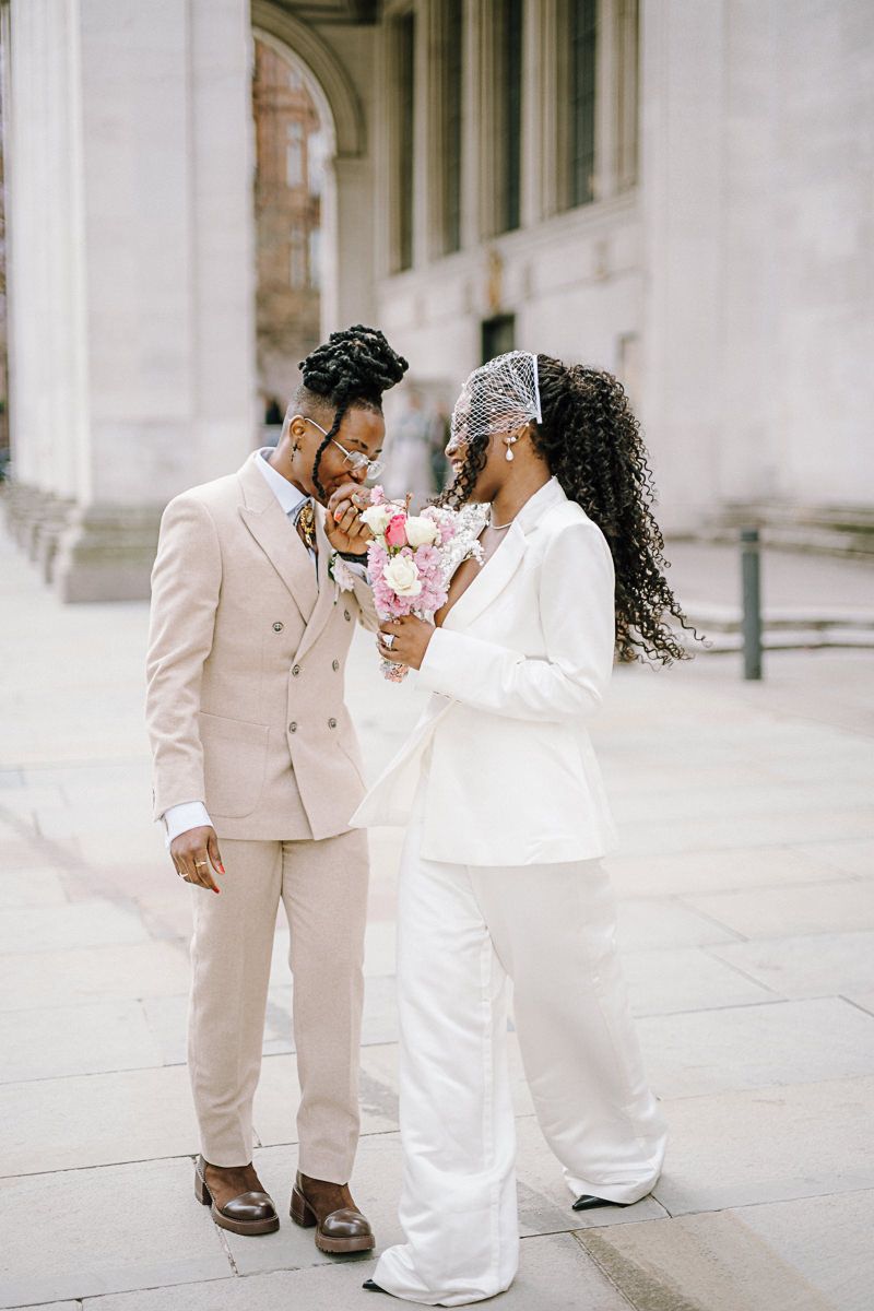 A couple in wedding attire, groom in beige suit, bride in white pantsuit, share a tender moment outdoors.