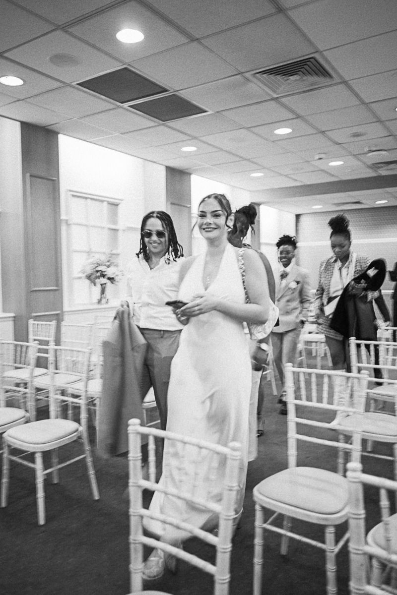 Smiling bride in white gown walks down aisle at indoor wedding ceremony with guests seated in chiavari chairs.