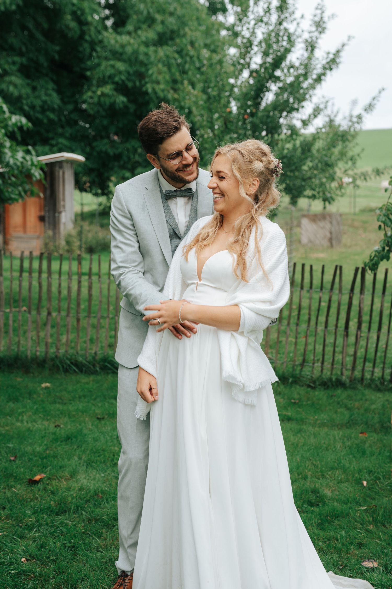 A couple in wedding attire stands together during golden hour in a scenic outdoor setting.