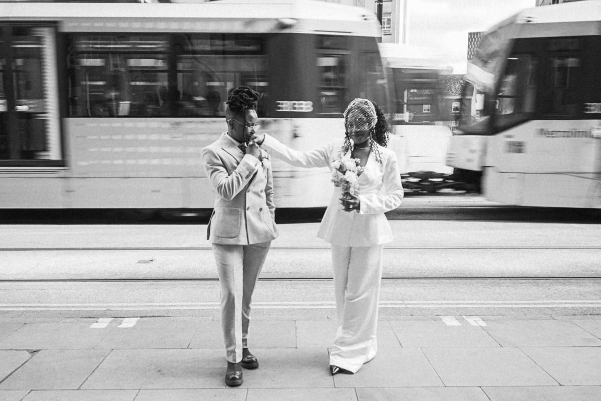 Black and white photo of two brides laughing in front of a passing tram on their wedding day.