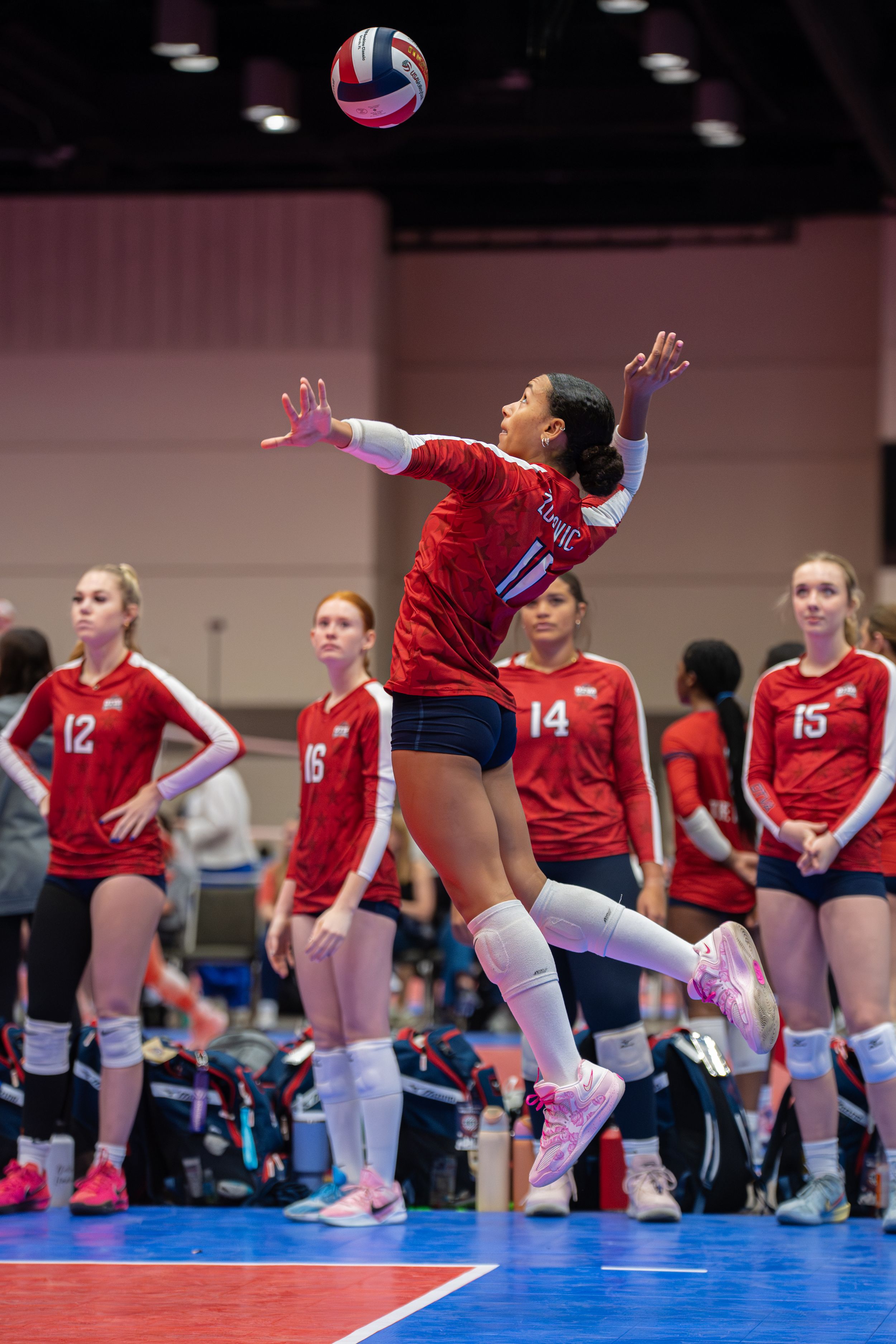 Female volleyball player in red jersey jumps to serve during indoor tournament while teammates watch from the sideline.