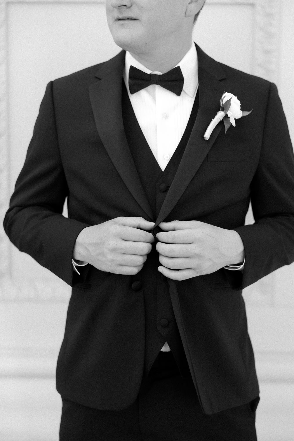 Groom in black tuxedo with bow tie and boutonniere adjusting jacket buttons, elegant black and white wedding photo.