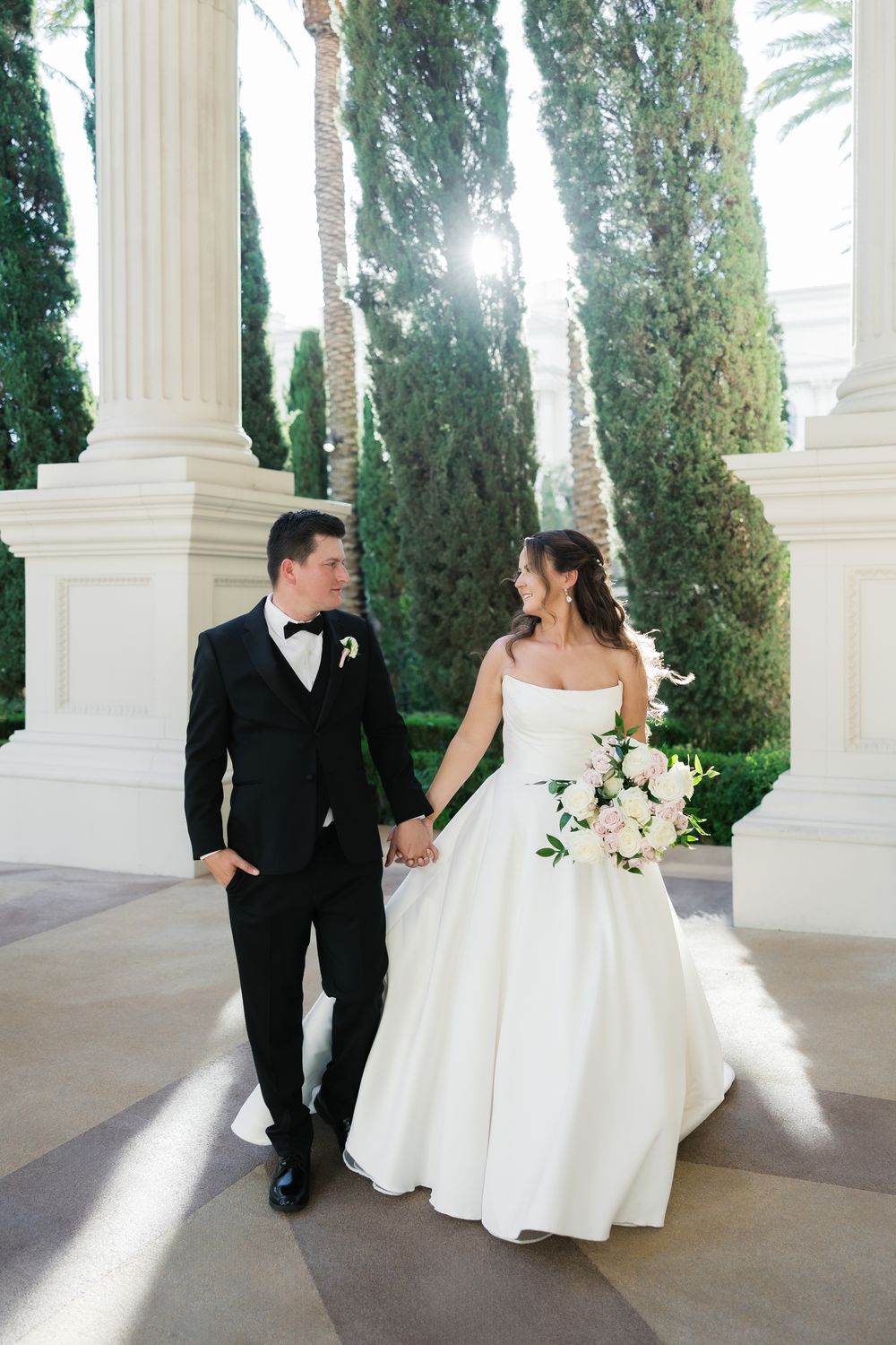 Bride and groom holding hands in sunlit garden with tall cypress trees and white columns at luxury venue.
