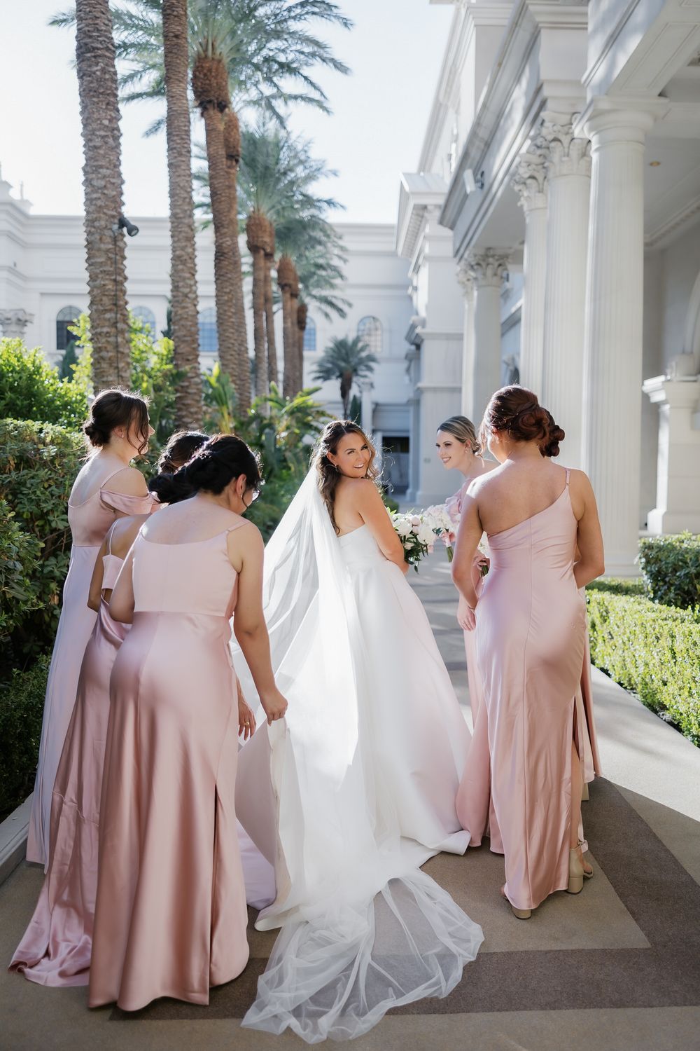 Bride in white gown and veil walks with bridesmaids in blush pink dresses outside elegant venue with palm trees.