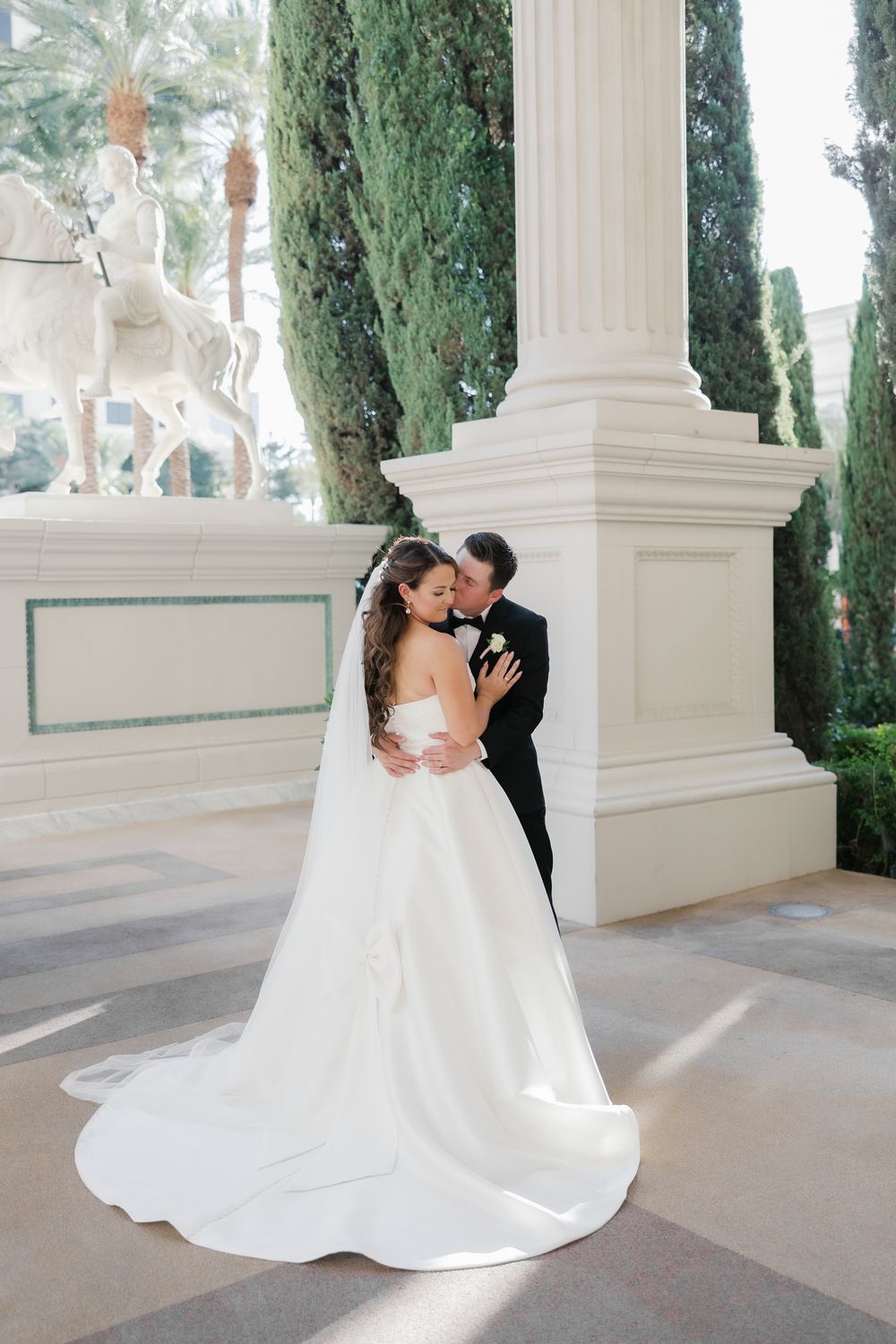 Groom embraces bride in stunning white gown near white columns with cypress trees in background.
